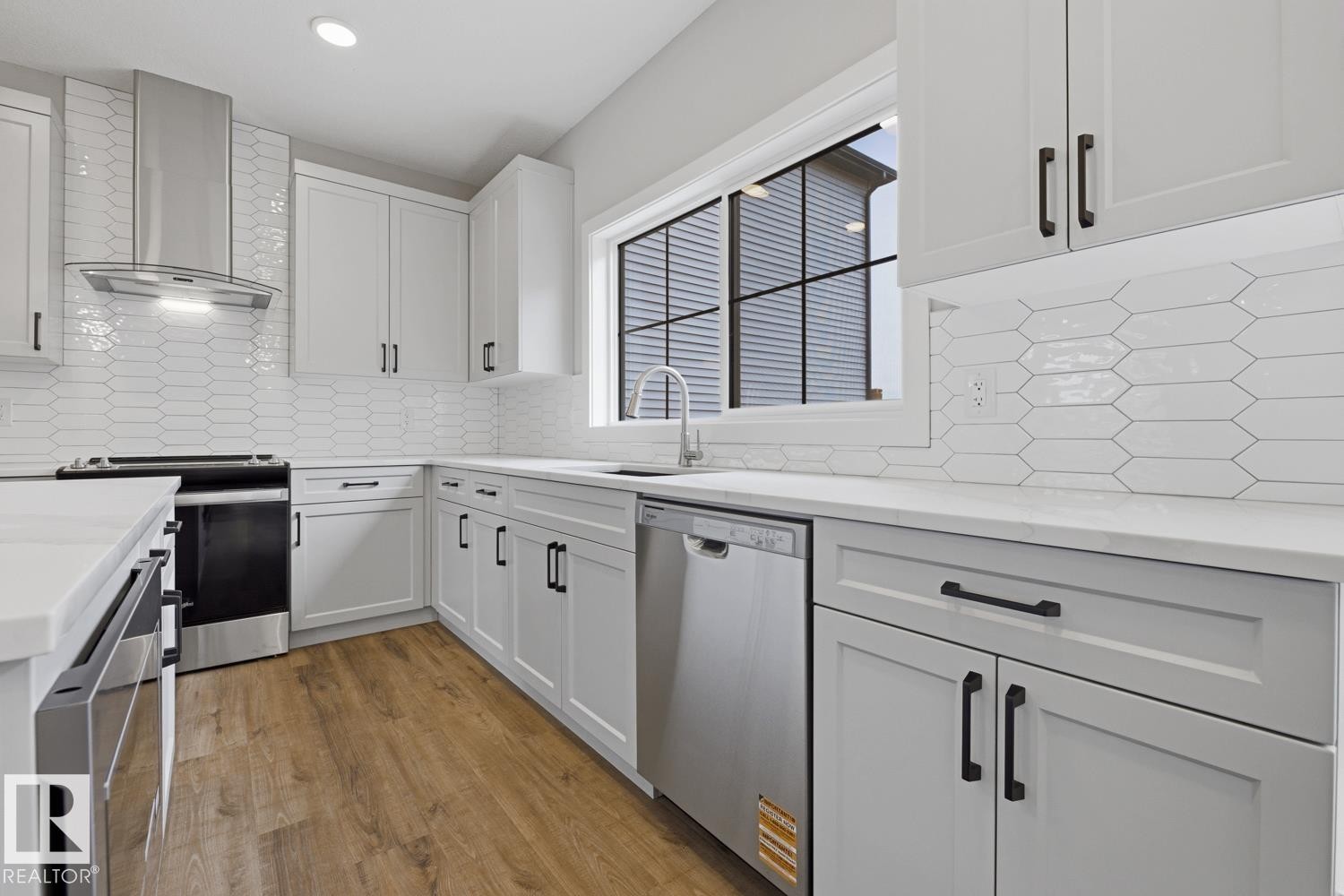Modern kitchen featuring white cabinetry with black hardware, stainless steel appliances, a geometric tile backsplash, light-toned countertops, and wood-finish flooring - 30 Chartres Close Nw, St. Albert, AB