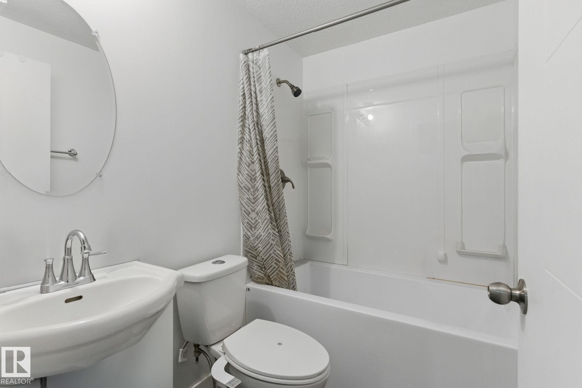 Bathroom featuring a pedestal sink with a chrome faucet, a round wall mirror, and a bathtub with a shower and a patterned shower curtain - 2416 Wonnacott Crest Sw, Edmonton, AB - Indoor Photo Showing Bathroom