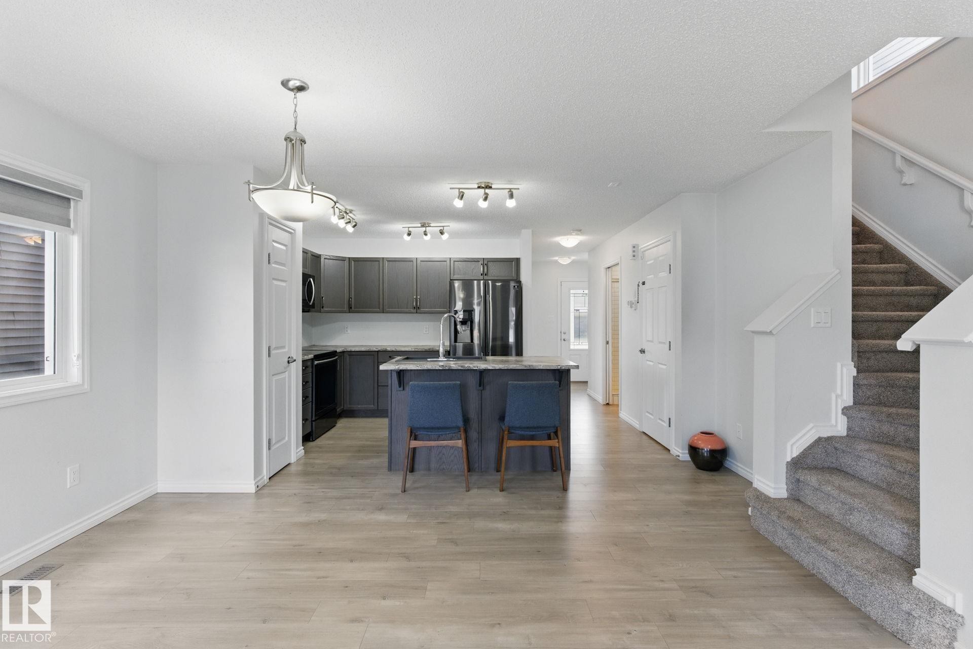 The open concept living area features light-toned flooring, a kitchen with dark cabinetry and stainless steel appliances, and a central island with seating - 2416 Wonnacott Crest Sw, Edmonton, AB - Indoor