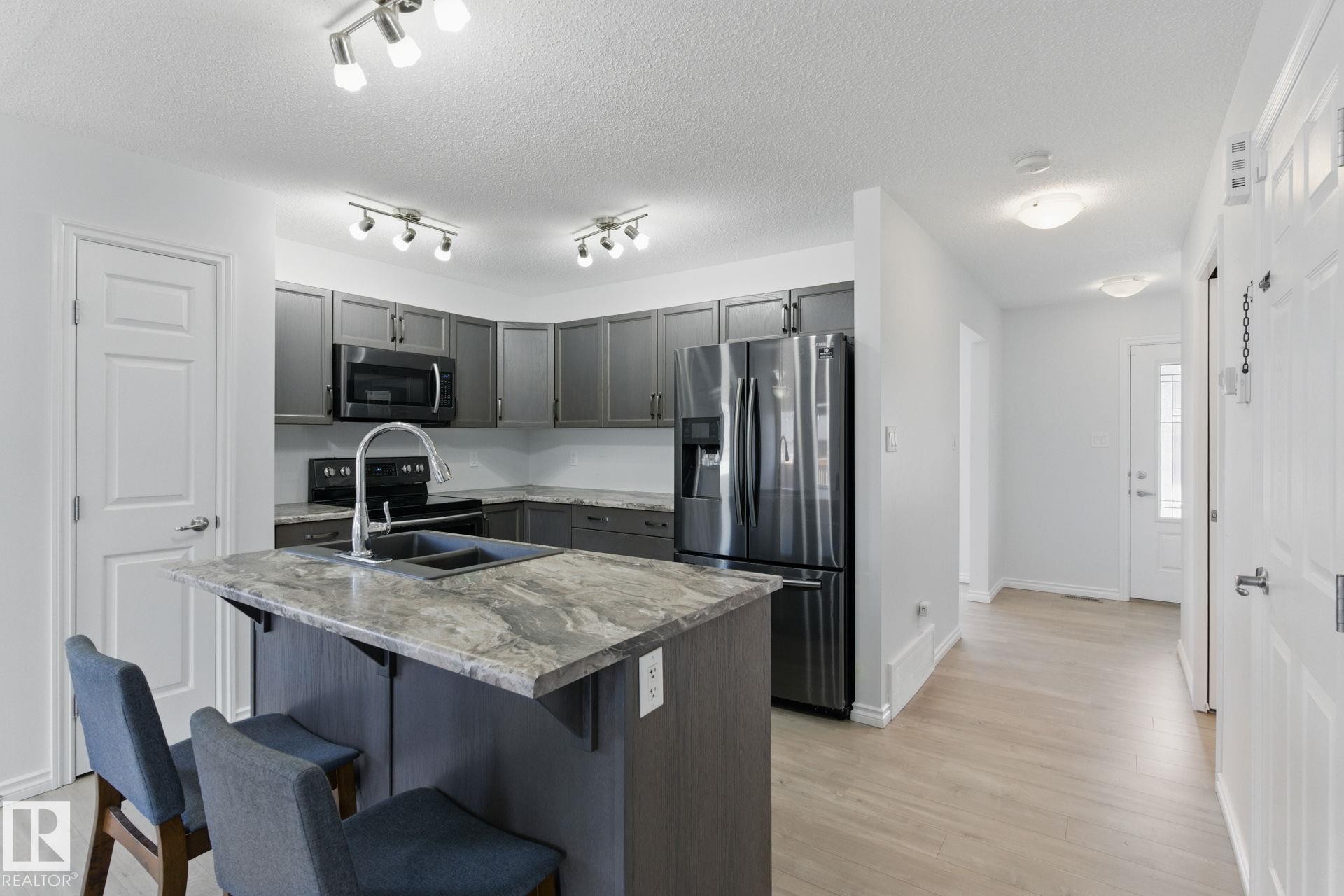 The kitchen features grey cabinetry, a kitchen island with a double sink, dark countertops, and stainless steel appliances - 2416 Wonnacott Crest Sw, Edmonton, AB - Indoor Photo Showing Kitchen With Double Sink