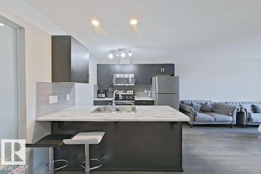 The kitchen features dark cabinetry, stainless steel appliances, a white marble-patterned countertop, and a double basin sink - 429 390 Windermere Rd, Edmonton, AB - Indoor Photo Showing Kitchen With Stainless Steel Kitchen With Double Sink With Upgraded Kitchen