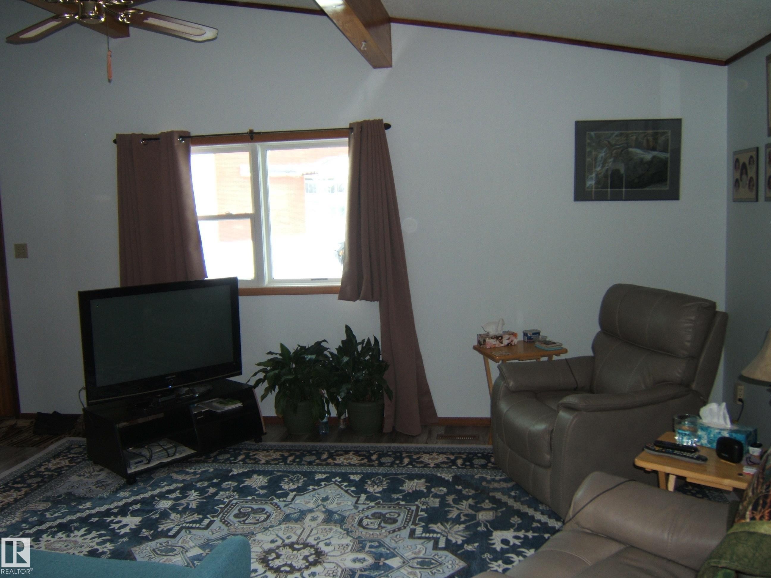The living area features a vaulted ceiling with exposed wooden beams and a ceiling fan, a window, and a patterned area rug - 4702 52 Avenue, Two Hills, AB - Indoor