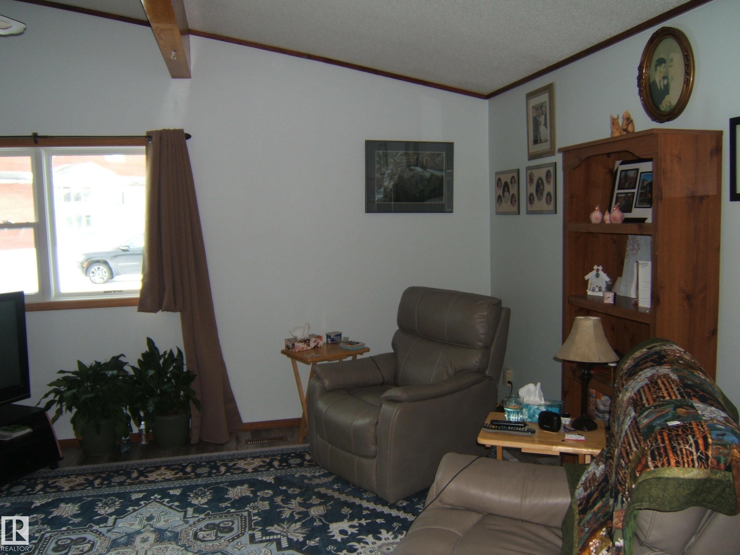 Living space featuring a vaulted ceiling with exposed beam, window with wood trim, and a patterned area rug - 4702 52 Avenue, Two Hills, AB - Indoor