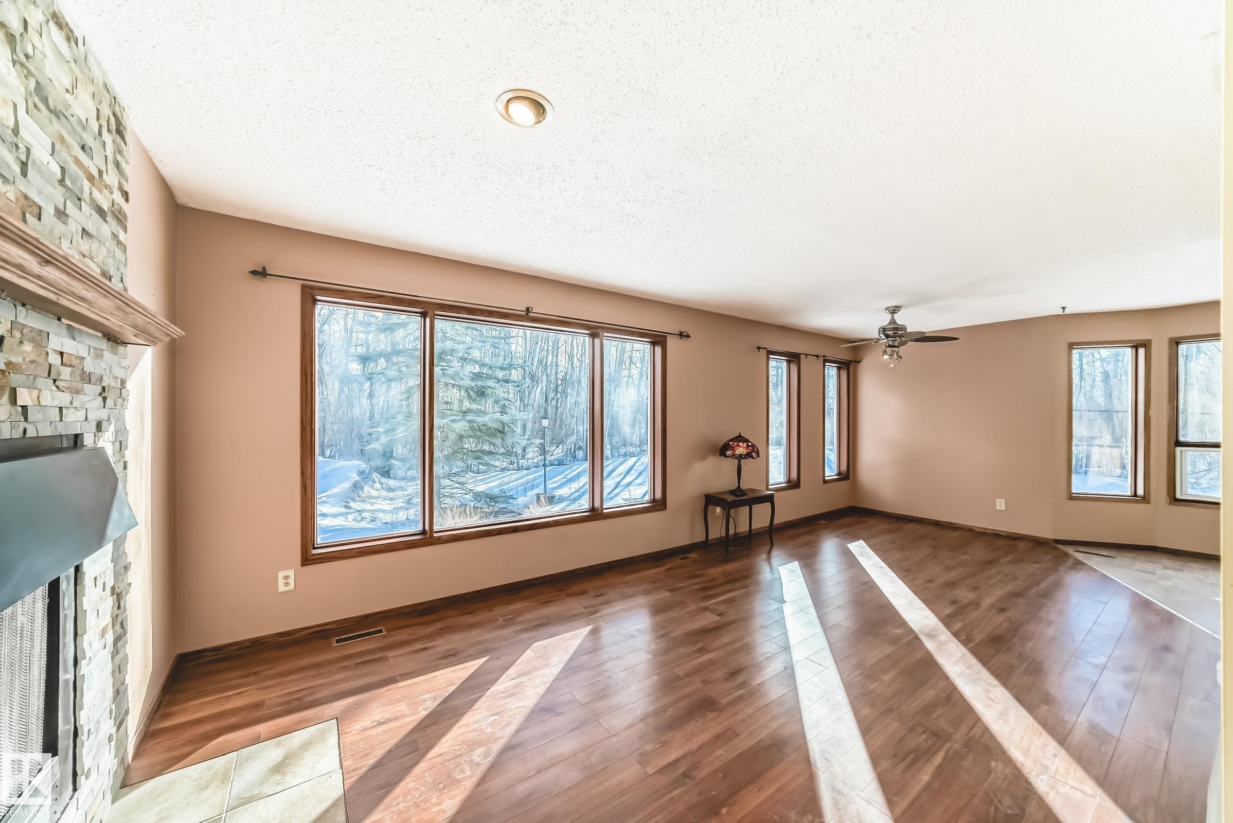 This living space features rich hardwood flooring, a stone-faced fireplace with a wooden mantel, and large windows that provide abundant natural light - 100 23016 Twp Rd 504, Rural Leduc County, AB - Indoor Photo Showing Other Room