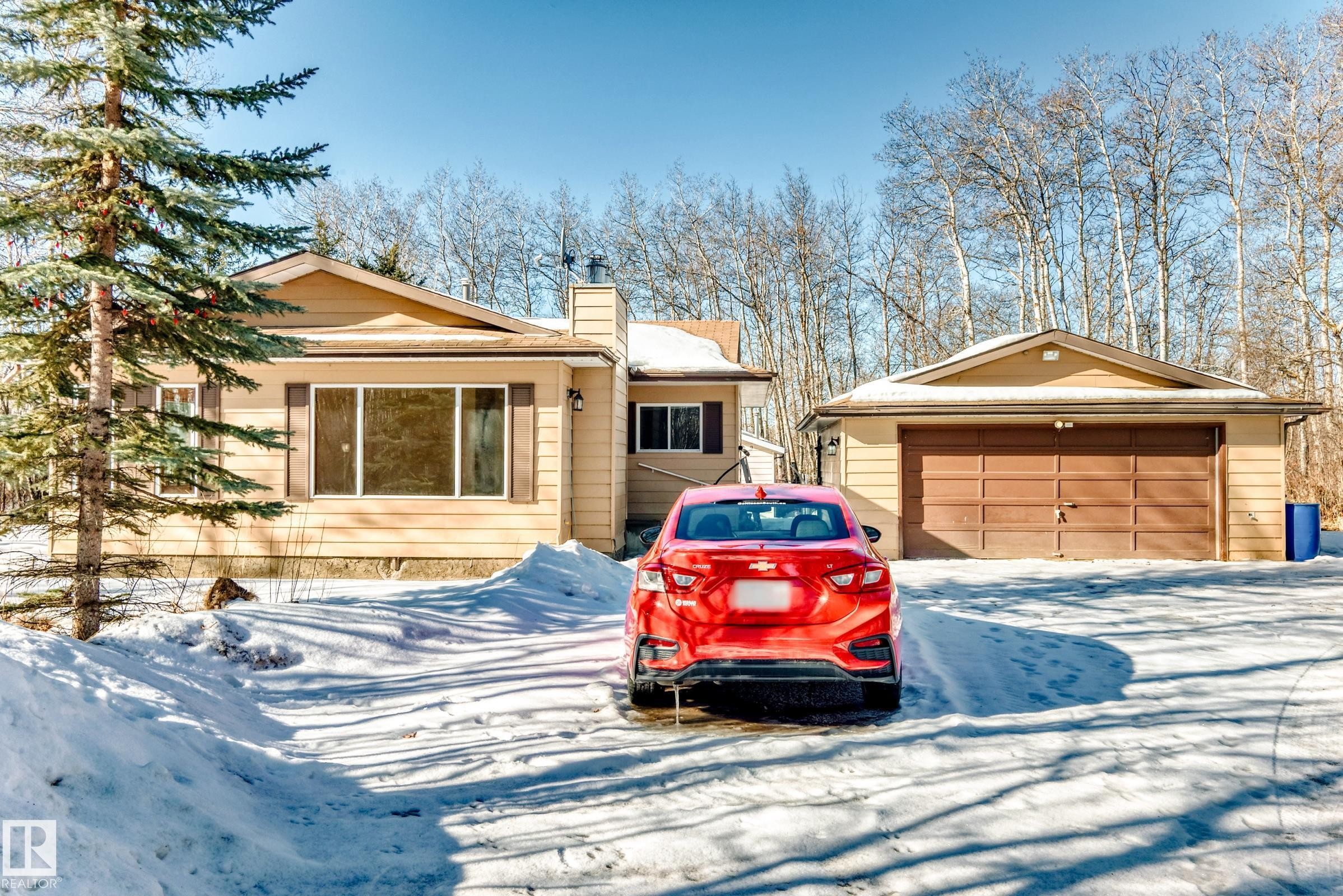 The property features a detached garage with a brown garage door, a light-colored exterior, and an expansive snow-covered driveway - 100 23016 Twp Rd 504, Rural Leduc County, AB - Outdoor