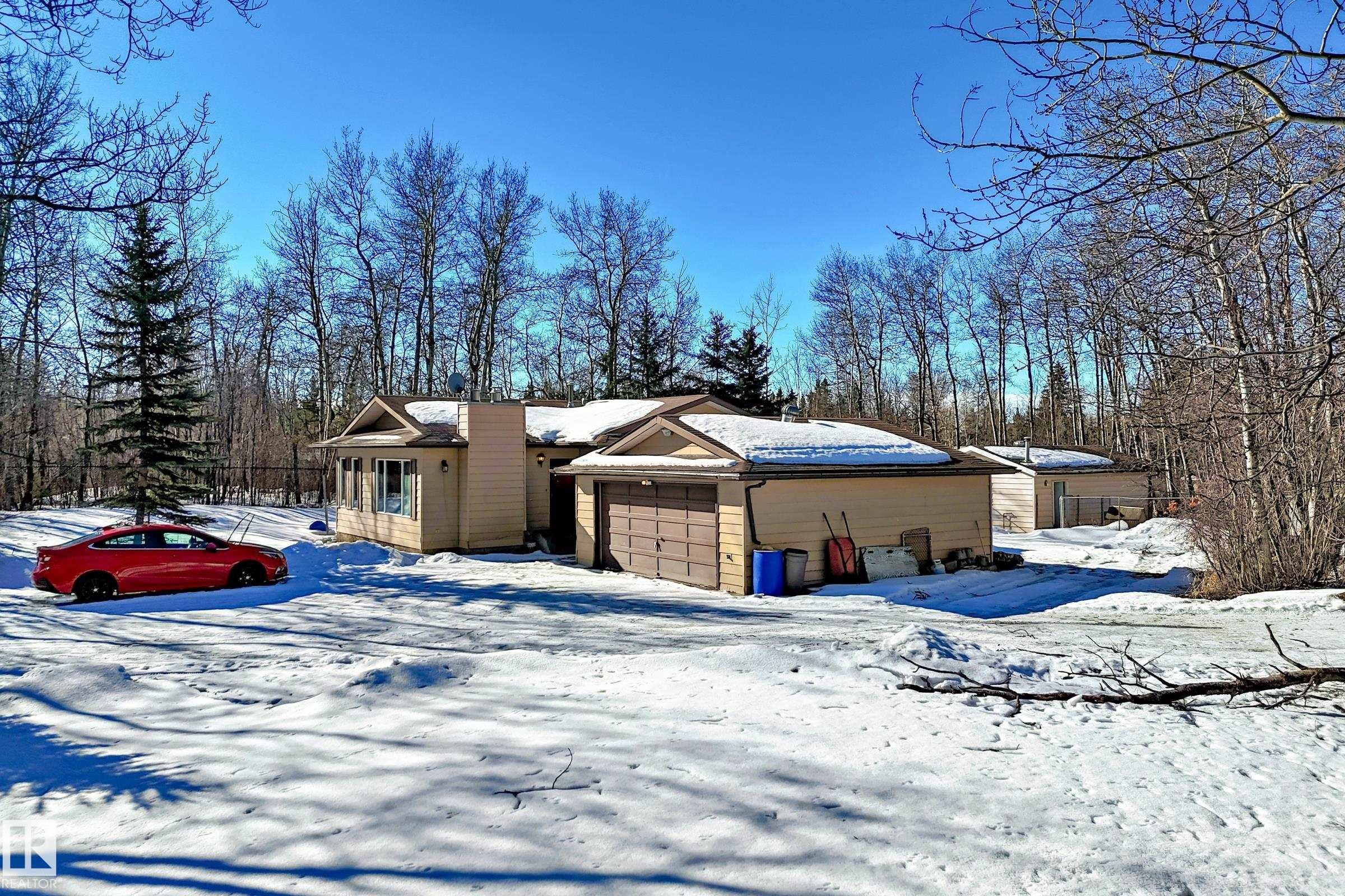 The property features a single-story structure with an attached garage and a detached outbuilding, set amidst a wooded backdrop - 100 23016 Twp Rd 504, Rural Leduc County, AB - Outdoor