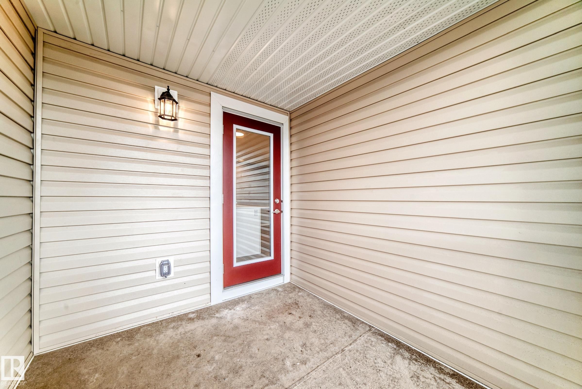 Private patio area featuring a red exterior door with full-light glass, beige horizontal siding, a concrete floor, and an exterior wall lantern - 20415 16 Avenue, Edmonton, AB