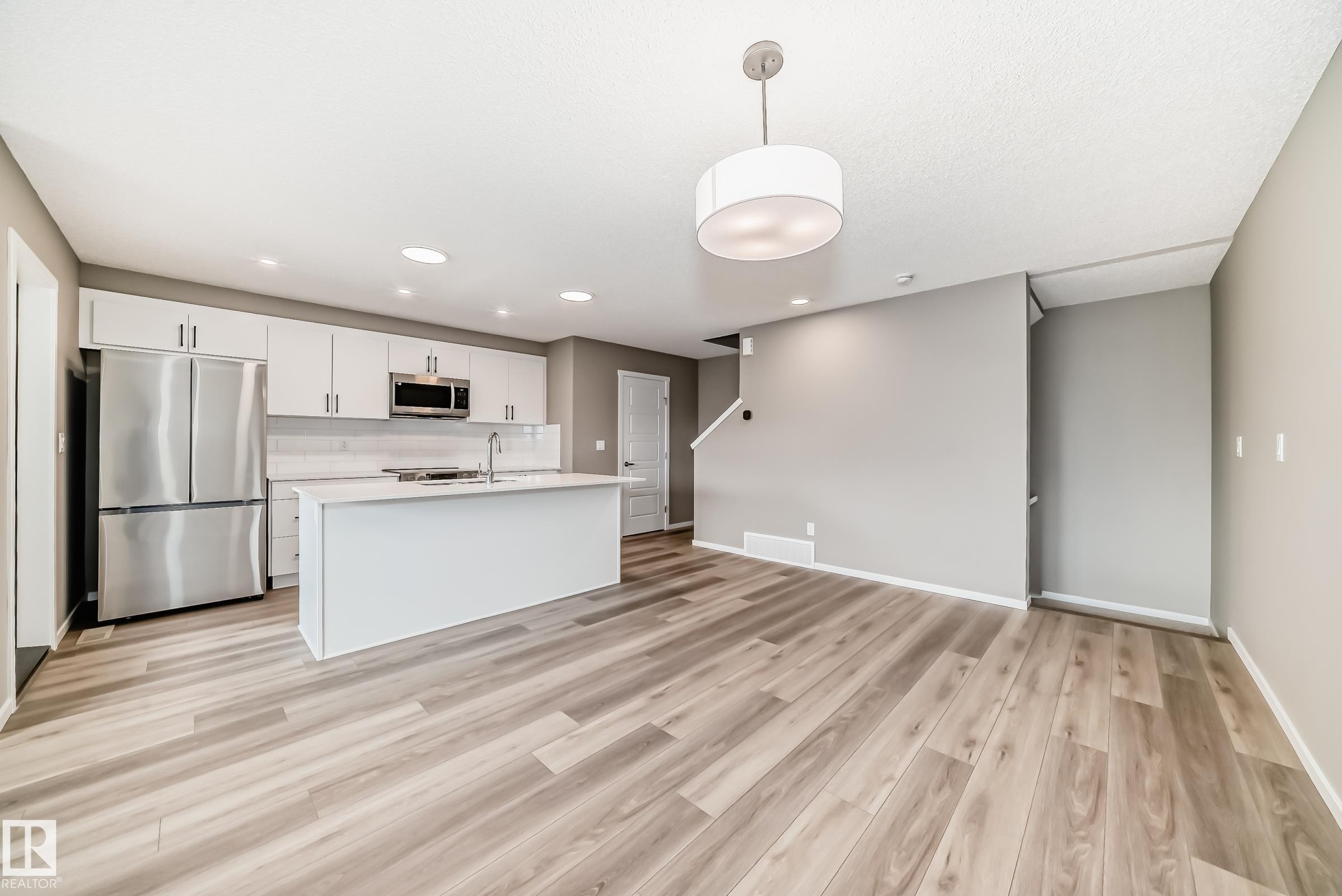 Open-concept living space featuring wood-finish flooring, a contemporary kitchen with white cabinetry, stainless steel appliances, a center island with a sink, and recessed lighting - 20415 16 Avenue, Edmonton, AB
