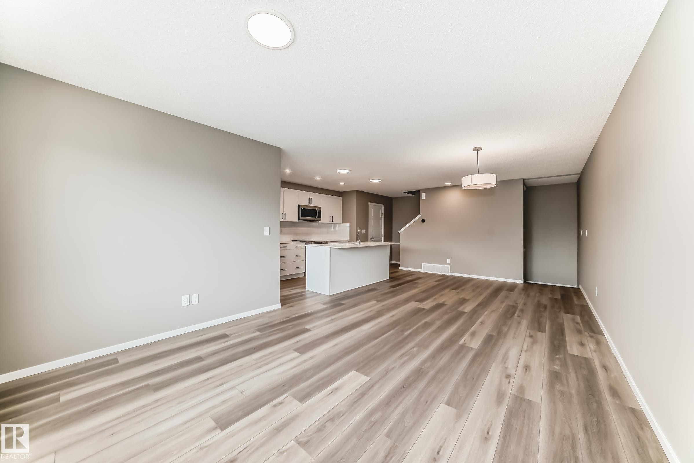 Expansive open-concept room featuring wood-finish flooring, a contemporary ceiling light fixture, recessed lighting, and a white baseboard trim - 20415 16 Avenue, Edmonton, AB