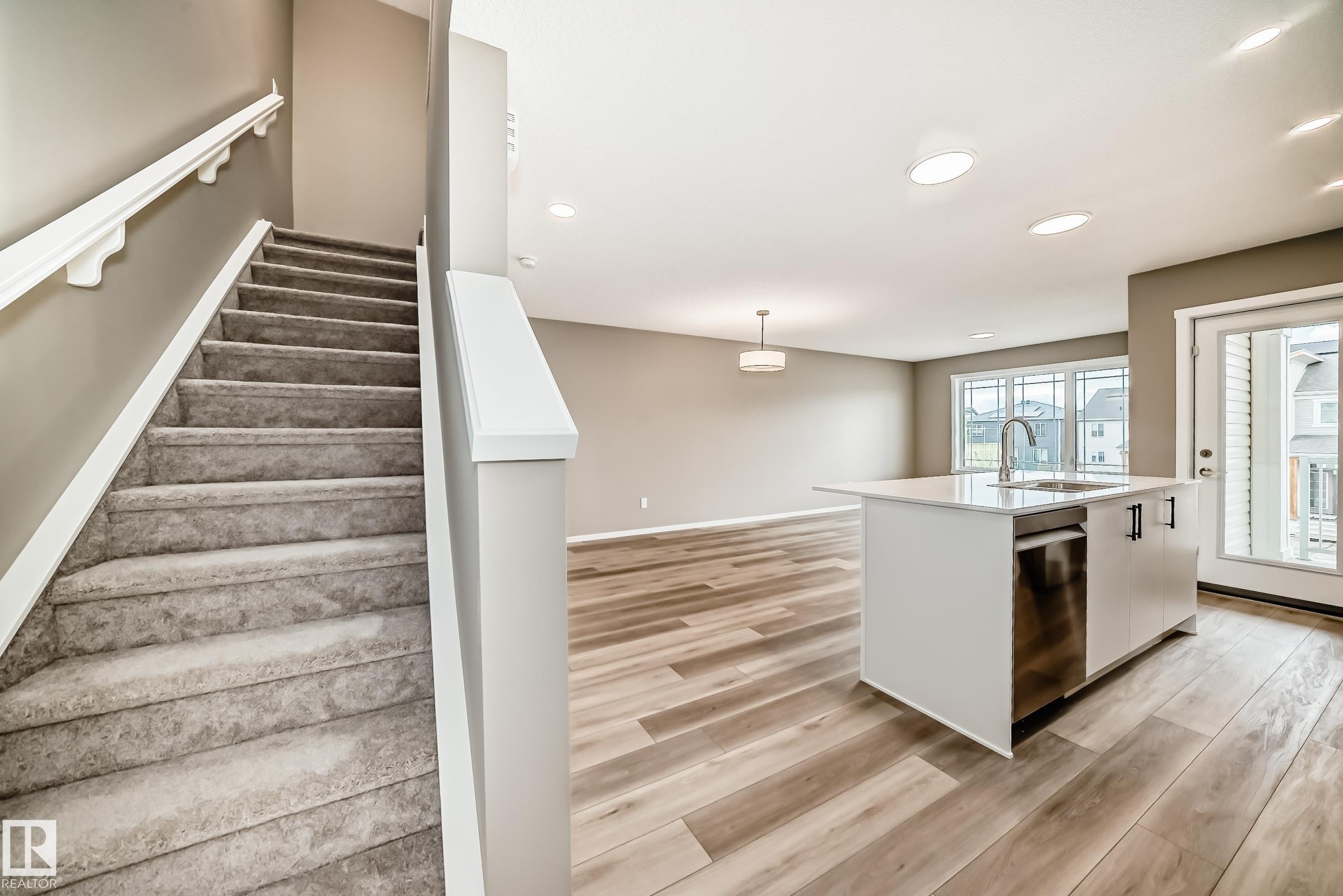 Open-concept living area featuring wood-finish flooring, a kitchen island with integrated sink, recessed lighting, and a glass-paneled door to an exterior balcony - 20415 16 Avenue, Edmonton, AB