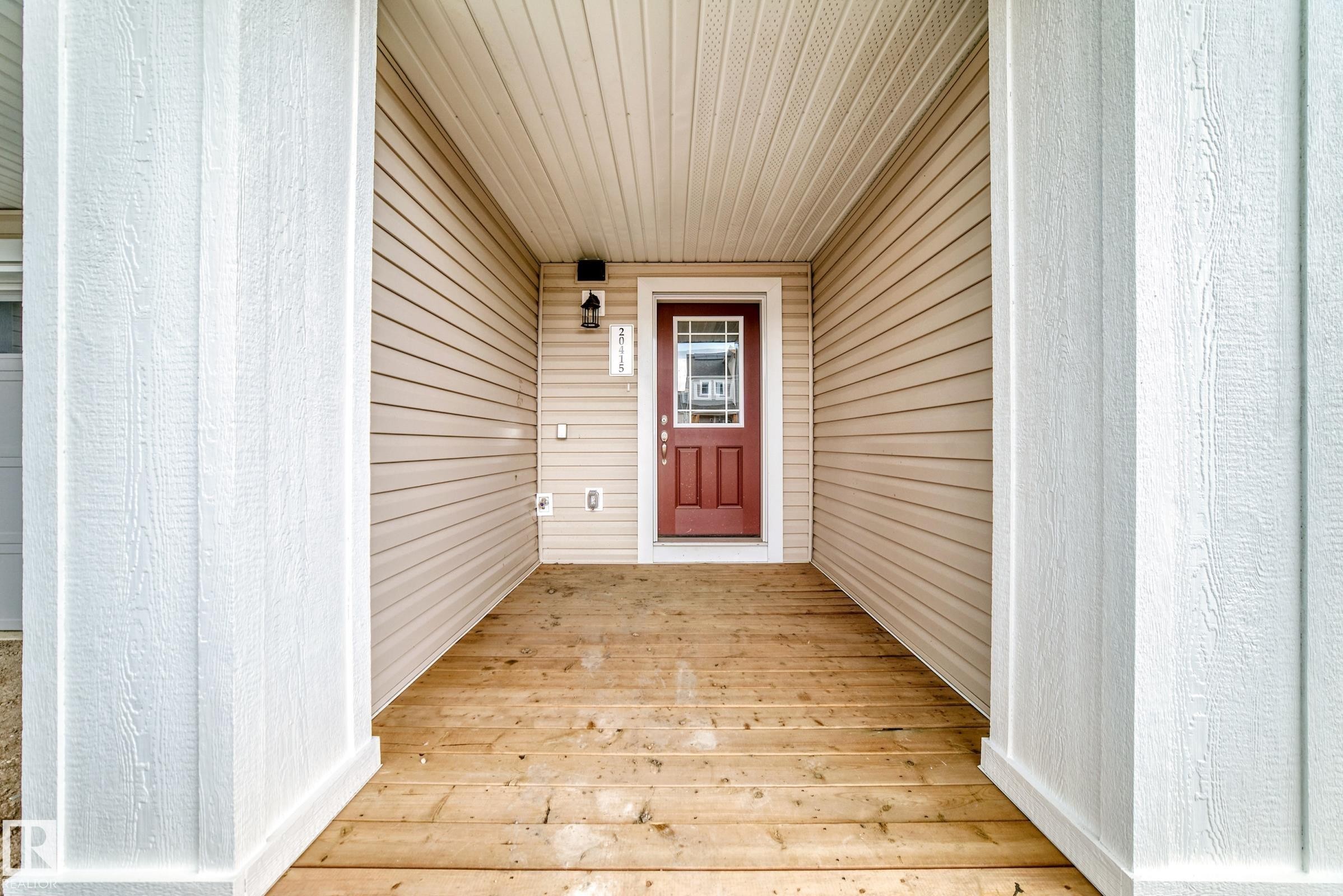 Covered front porch featuring wood-finish flooring, white architectural columns, beige vinyl siding, and a red entry door with decorative glass - 20415 16 Avenue, Edmonton, AB