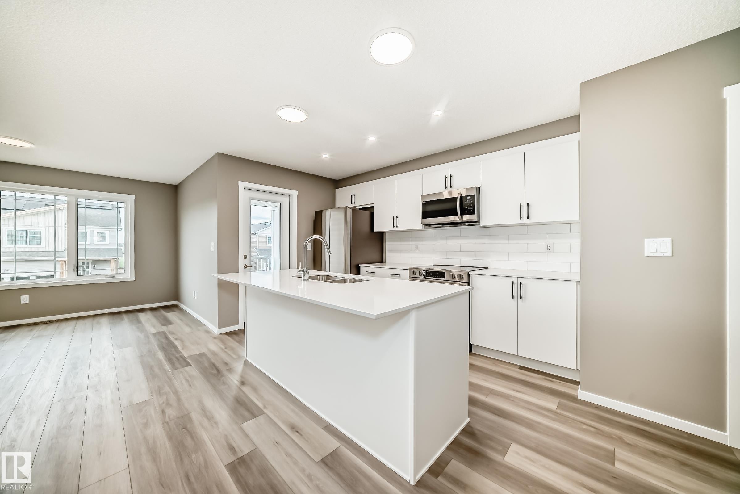 Modern kitchen featuring a central island with a sink, stainless steel appliances, white cabinetry, a subway tile backsplash, and wood-finish flooring - 20415 16 Avenue, Edmonton, AB