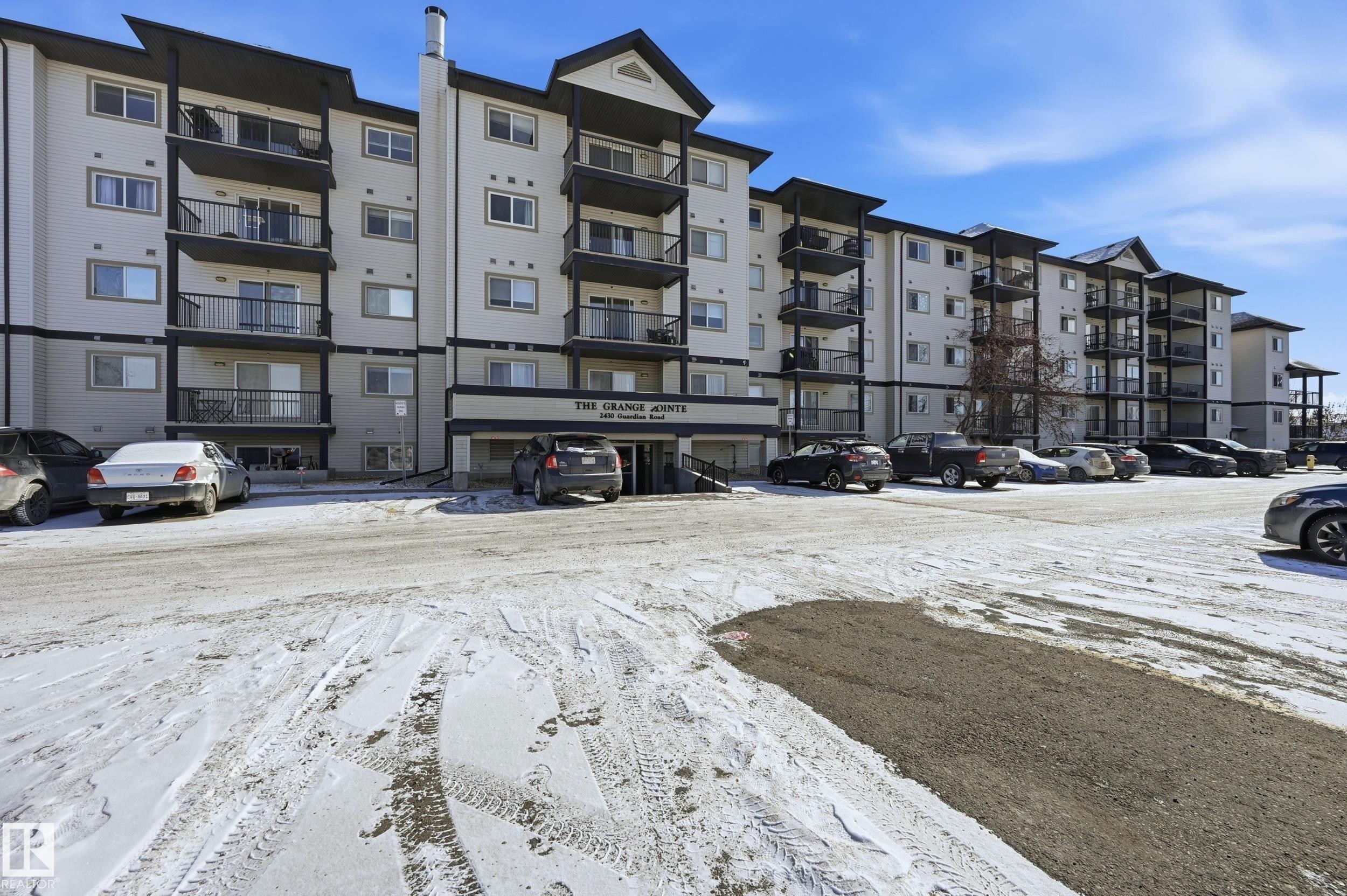 The property features a multi-story building with balconies, a main entrance with a canopy, and an expansive parking lot - 314 2430 Guardian Road, Edmonton, AB - Outdoor With Balcony With Facade
