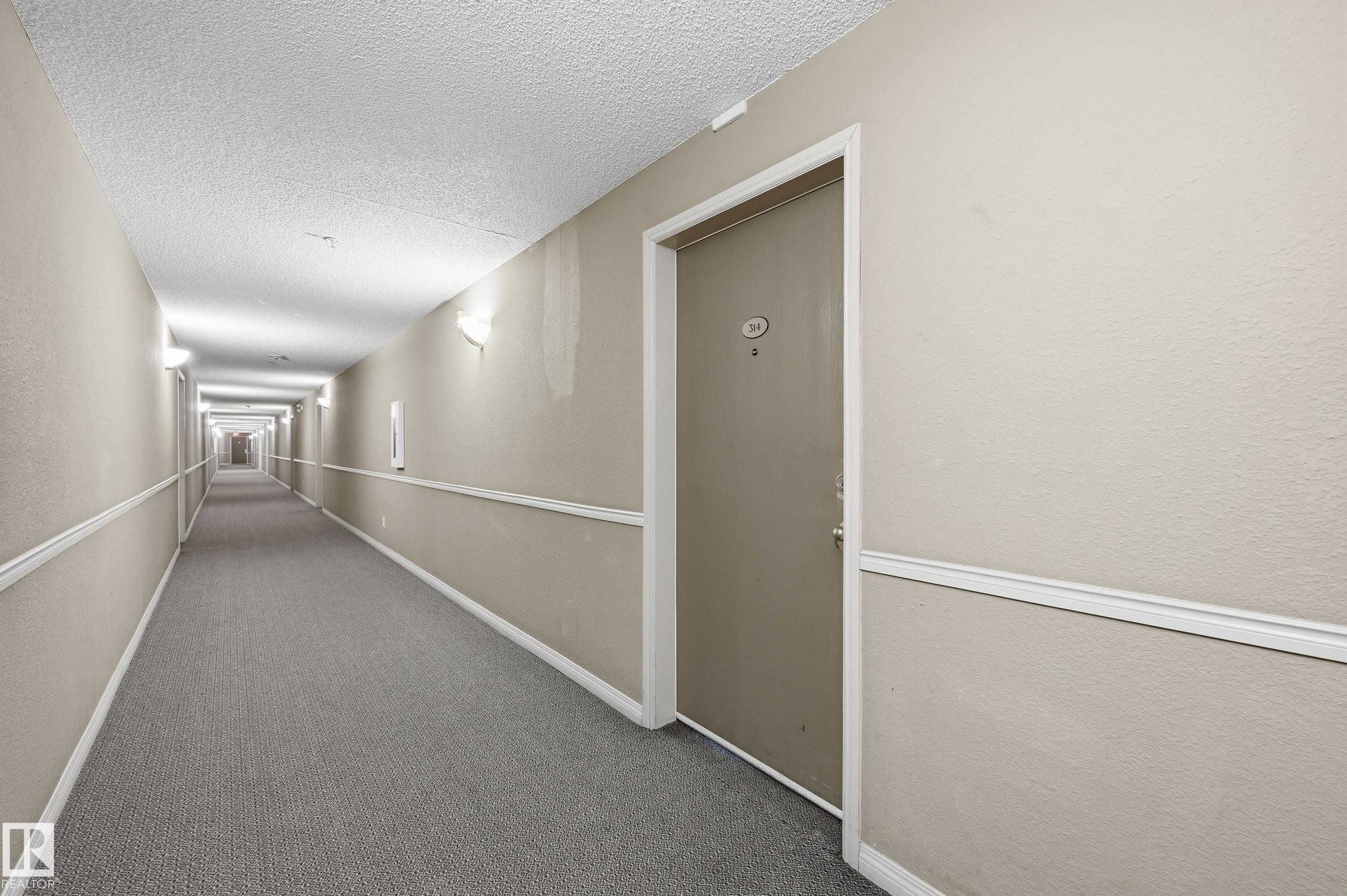 The property features a private entrance with a numbered door, set within a carpeted hallway with sconce lighting and crown molding details - 314 2430 Guardian Road, Edmonton, AB - Indoor Photo Showing Other Room