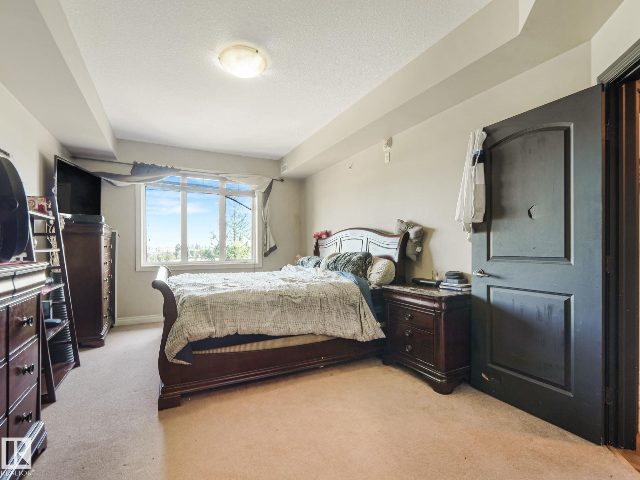 Bedroom featuring carpeted flooring, a large window providing natural light, and a dark wood door - 412 12408 15 Avenue, Edmonton, AB - Indoor Photo Showing Bedroom