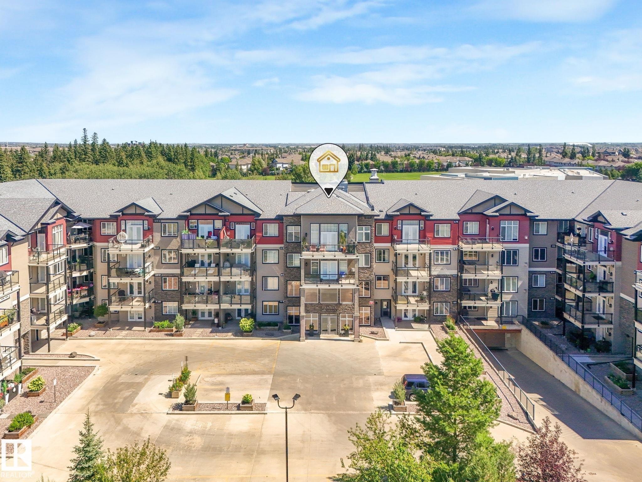 The property features a multi-story building with a red and grey exterior, balconies, and a paved parking area - 412 12408 15 Avenue, Edmonton, AB - Outdoor With Balcony With Facade