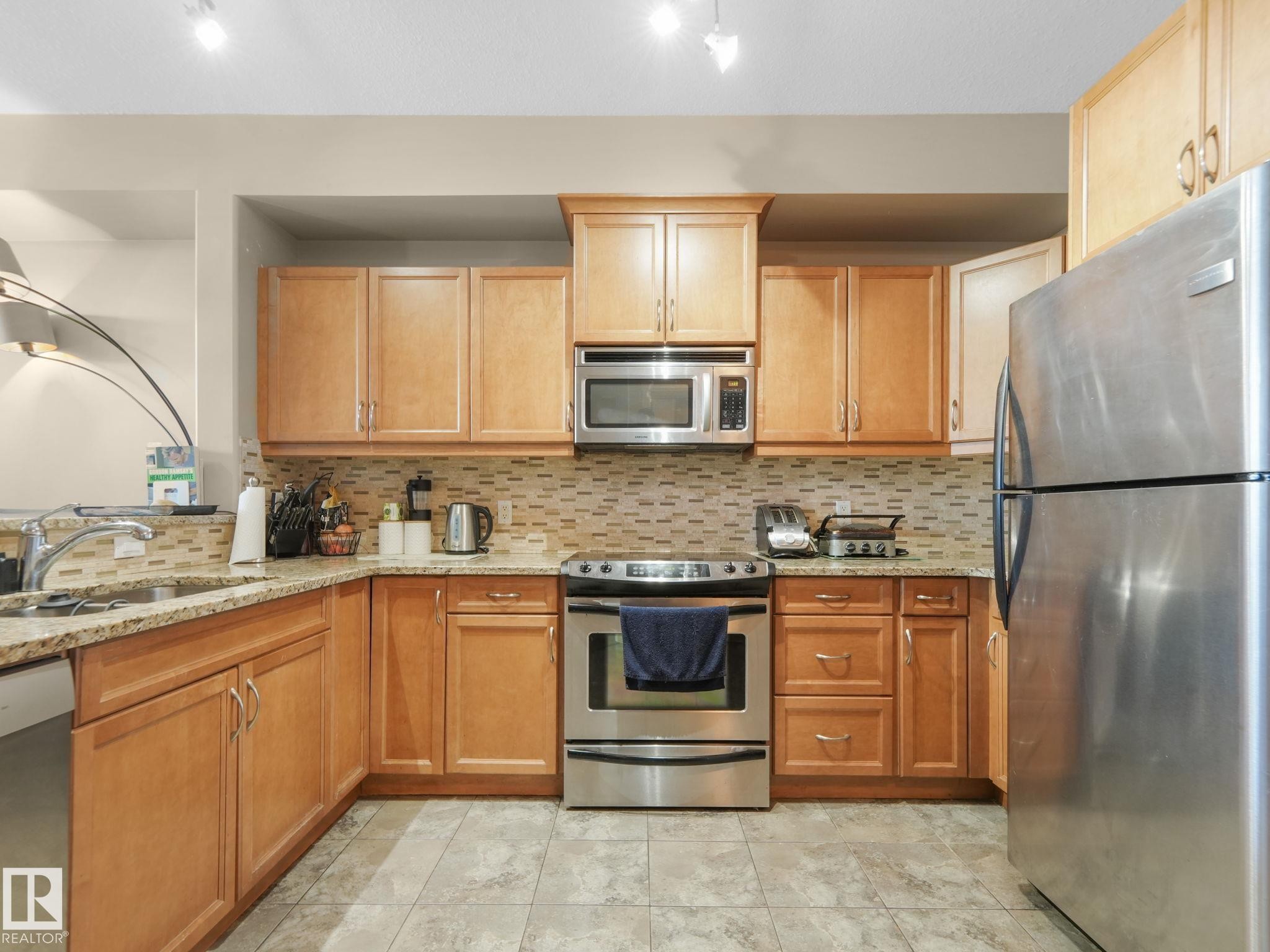 The kitchen features wood cabinetry, granite countertops, a tiled backsplash, stainless steel appliances, and tiled flooring - 412 12408 15 Avenue, Edmonton, AB - Indoor Photo Showing Kitchen