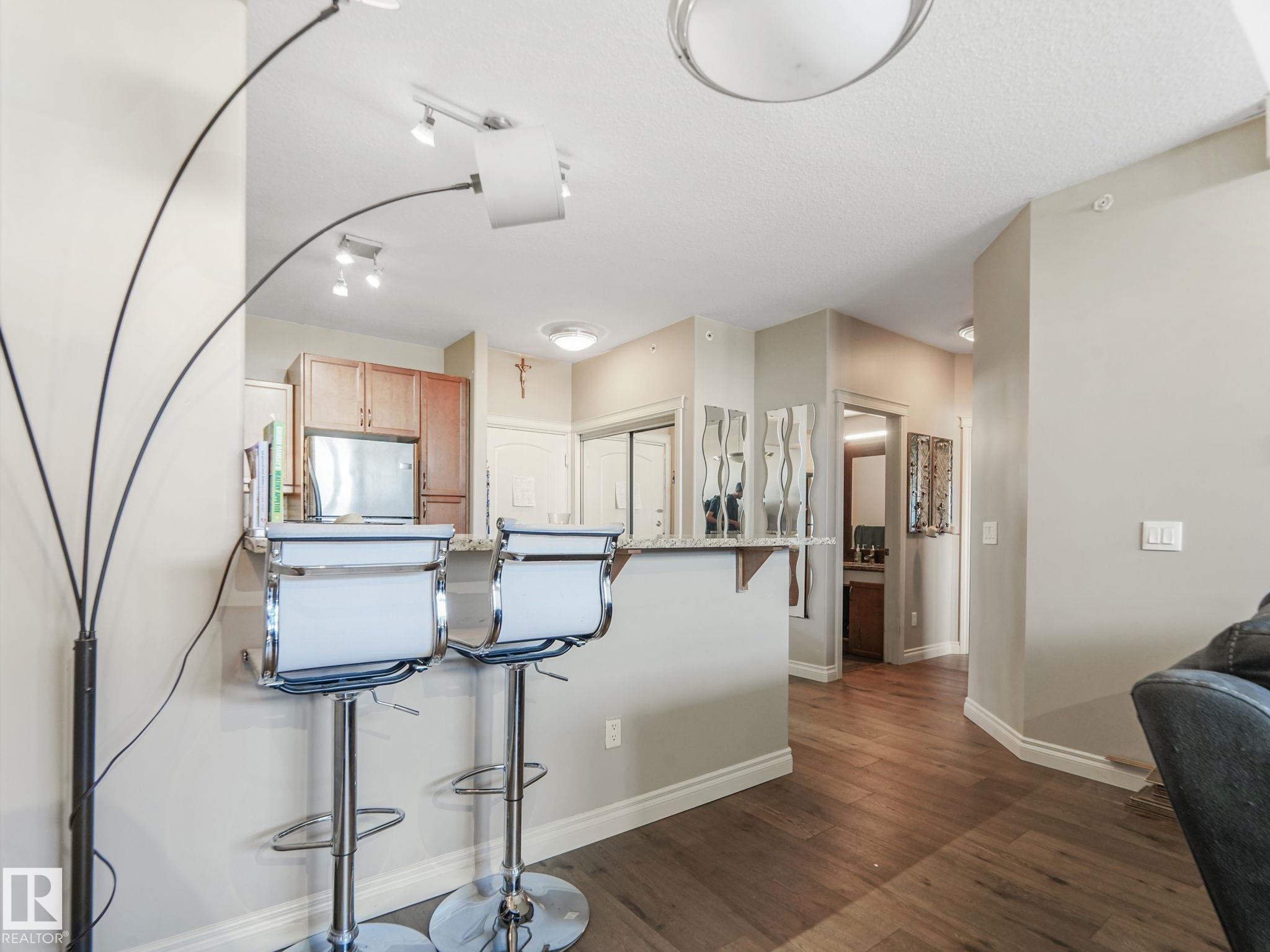 The property features hardwood floors, a kitchen area with wooden cabinetry and a stainless steel appliance, and a breakfast bar with seating - 412 12408 15 Avenue, Edmonton, AB - Indoor