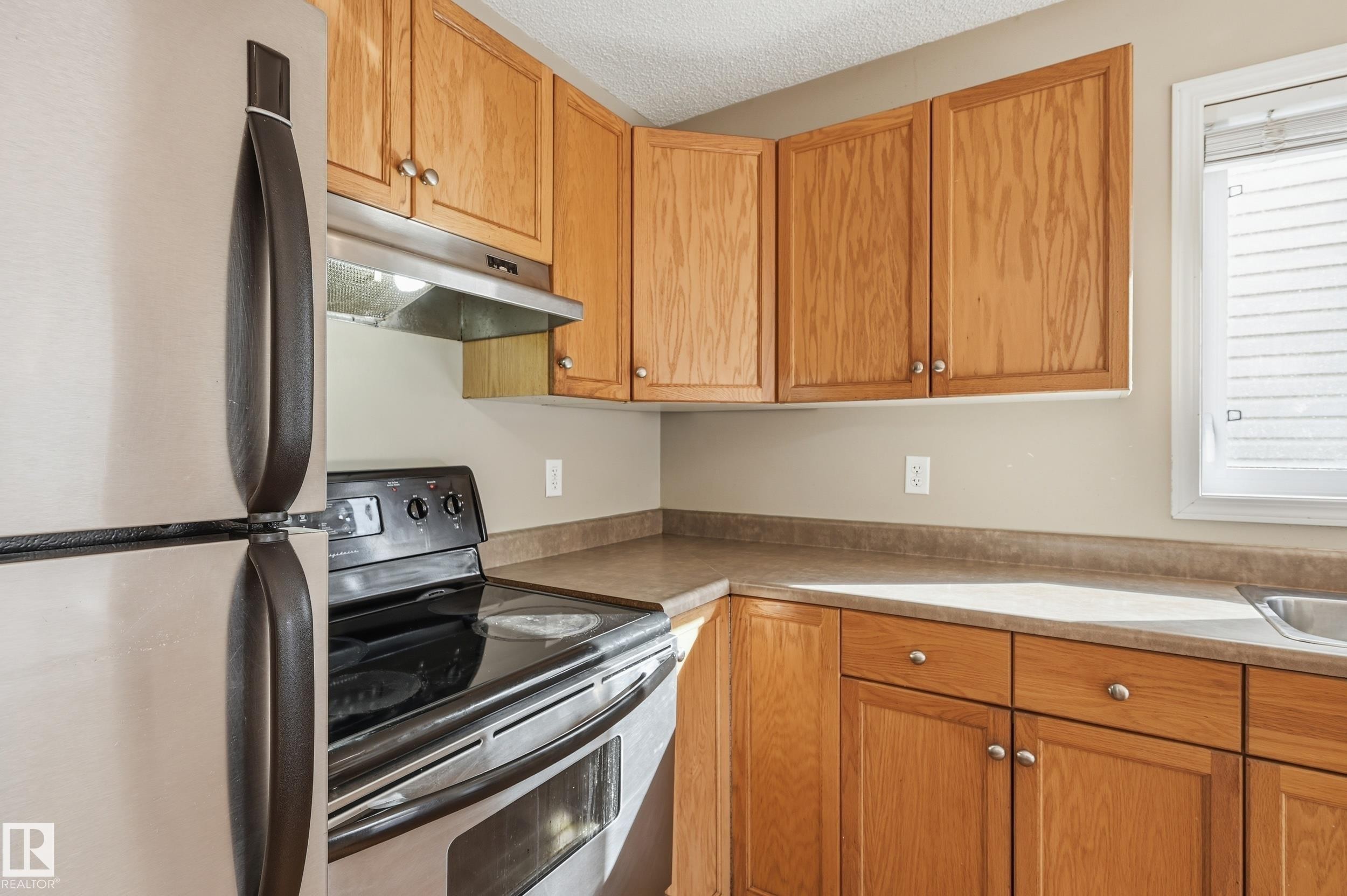 The kitchen features light wood cabinetry, a stainless steel refrigerator, and a stainless steel oven with a black cooktop - 53 287 Macewan Road, Edmonton, AB - Indoor Photo Showing Kitchen