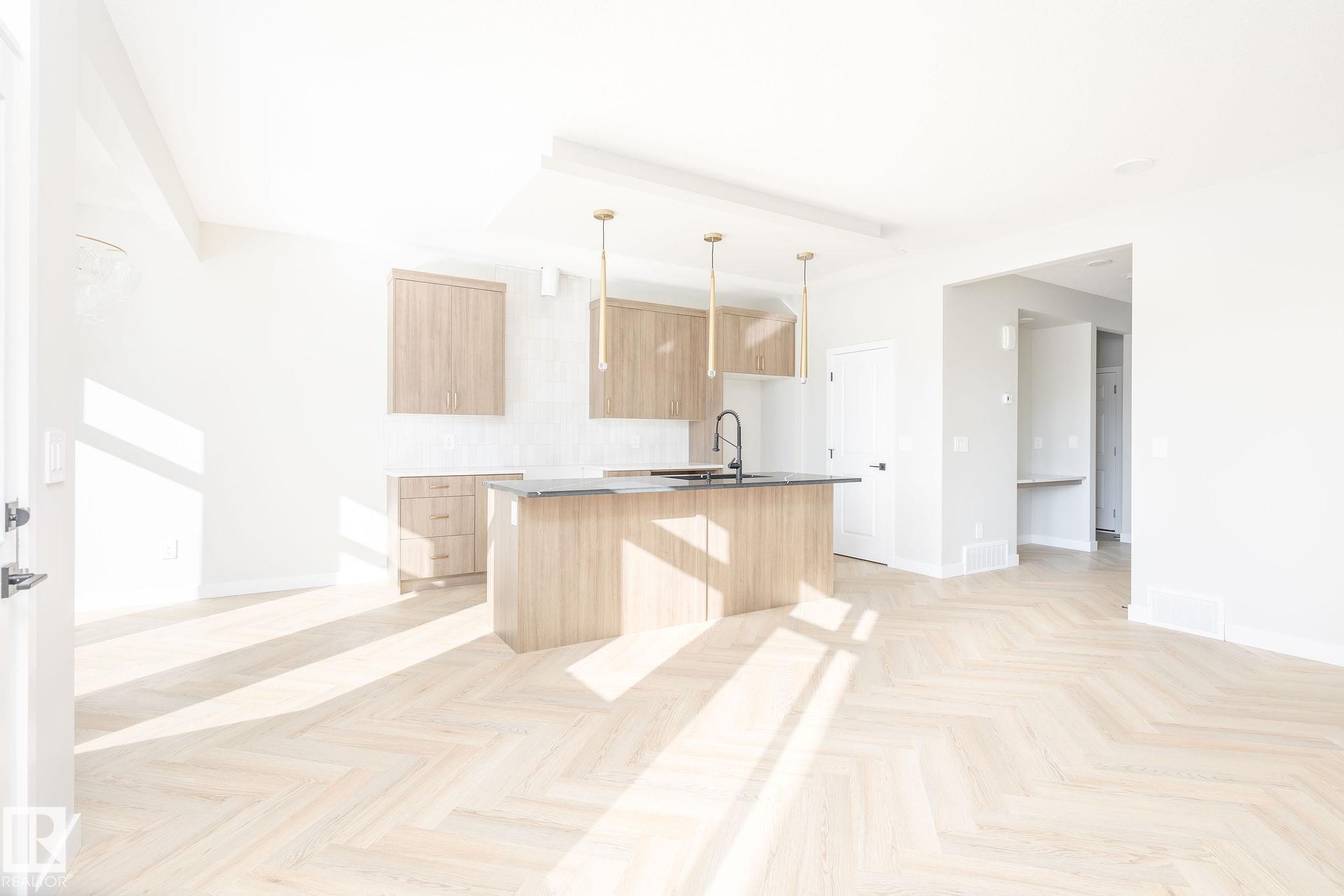 The property features light-toned hardwood flooring laid in a herringbone pattern, an open-concept kitchen with light wood cabinetry, a central island with a dark countertop and a sink, and modern pendant lighting fixtures - 2206 Crossbill Link, Edmonton, AB - Indoor