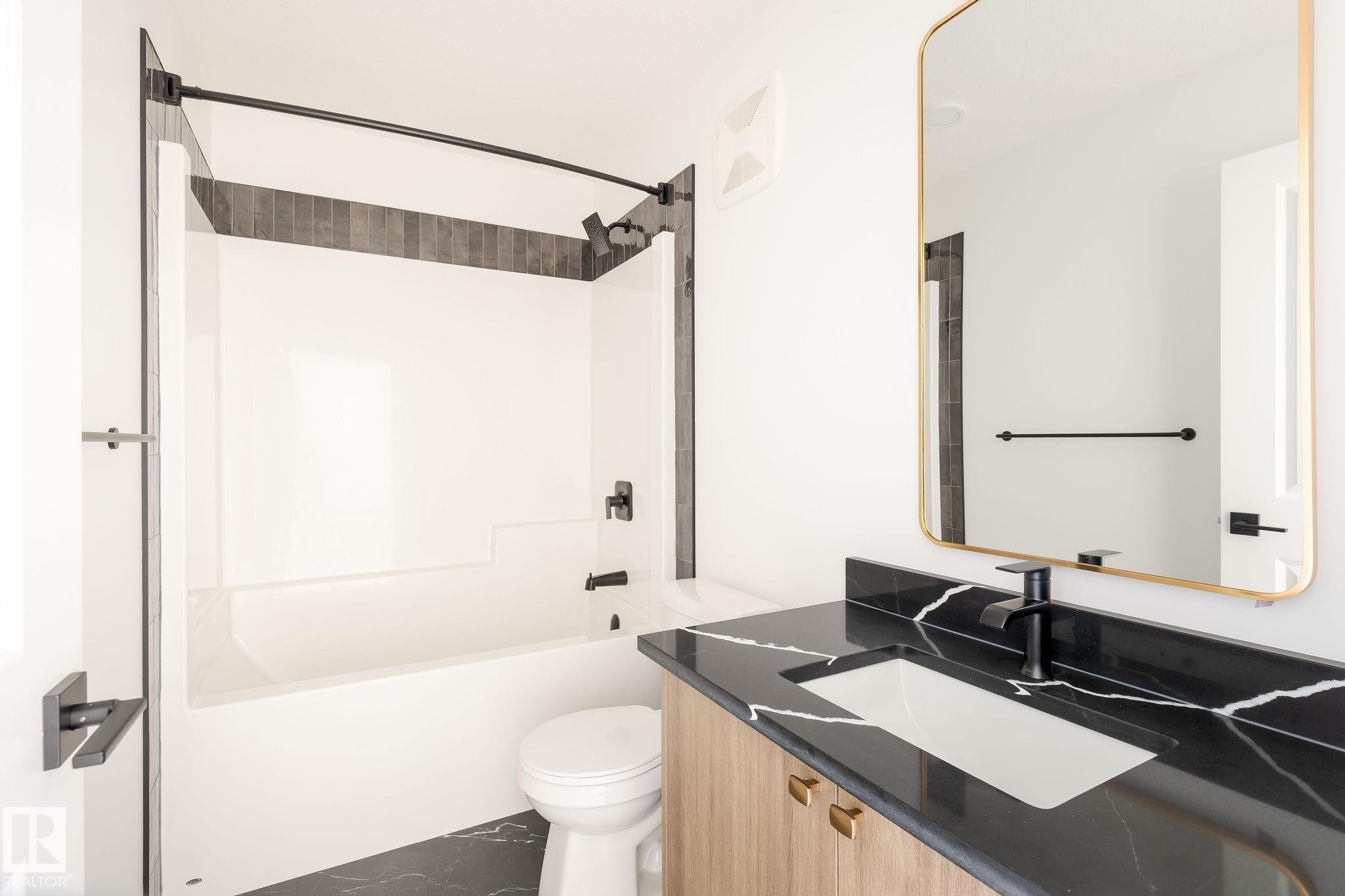 Bathroom featuring a white bathtub with dark accent tiles, a vanity with a black countertop and integrated sink, and a rectangular mirror with a gold-toned frame - 2206 Crossbill Link, Edmonton, AB - Indoor Photo Showing Bathroom