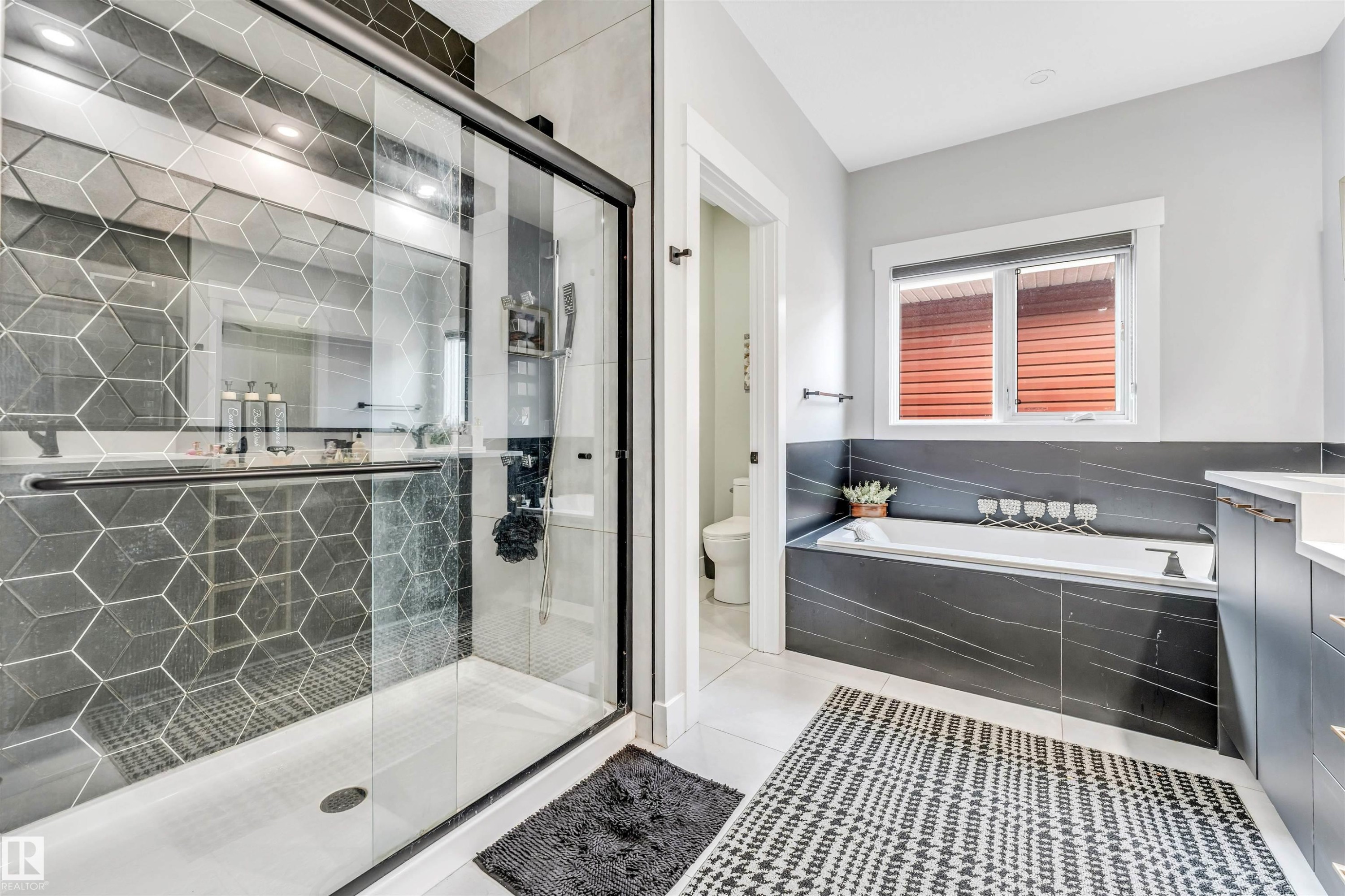 Well-appointed bathroom featuring a glass-enclosed shower with geometric tile, a built-in bathtub with dark stone-look surround, and a window with blinds - 1531 151 Avenue, Edmonton, AB - Indoor Photo Showing Bathroom