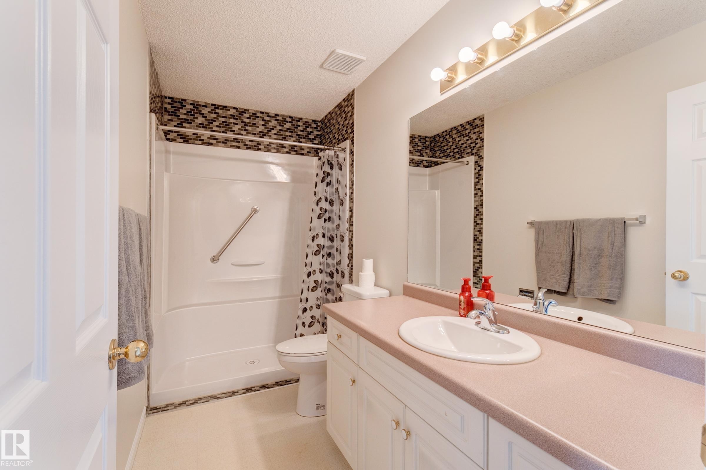 Bathroom featuring a shower with a grab bar, mosaic tile detailing, a vanity with an integrated sink, and a large mirror with overhead lighting - 5 308 Jackson Road, Edmonton, AB - Indoor Photo Showing Bathroom