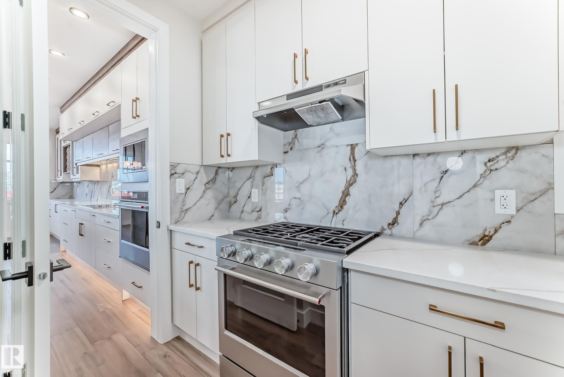 The kitchen features white cabinetry with gold-toned hardware, a stainless steel range with a ventilation hood, and marble-patterned backsplash and countertops - 7665 Kimiwan Crescent, Edmonton, AB - Indoor Photo Showing Kitchen With Upgraded Kitchen
