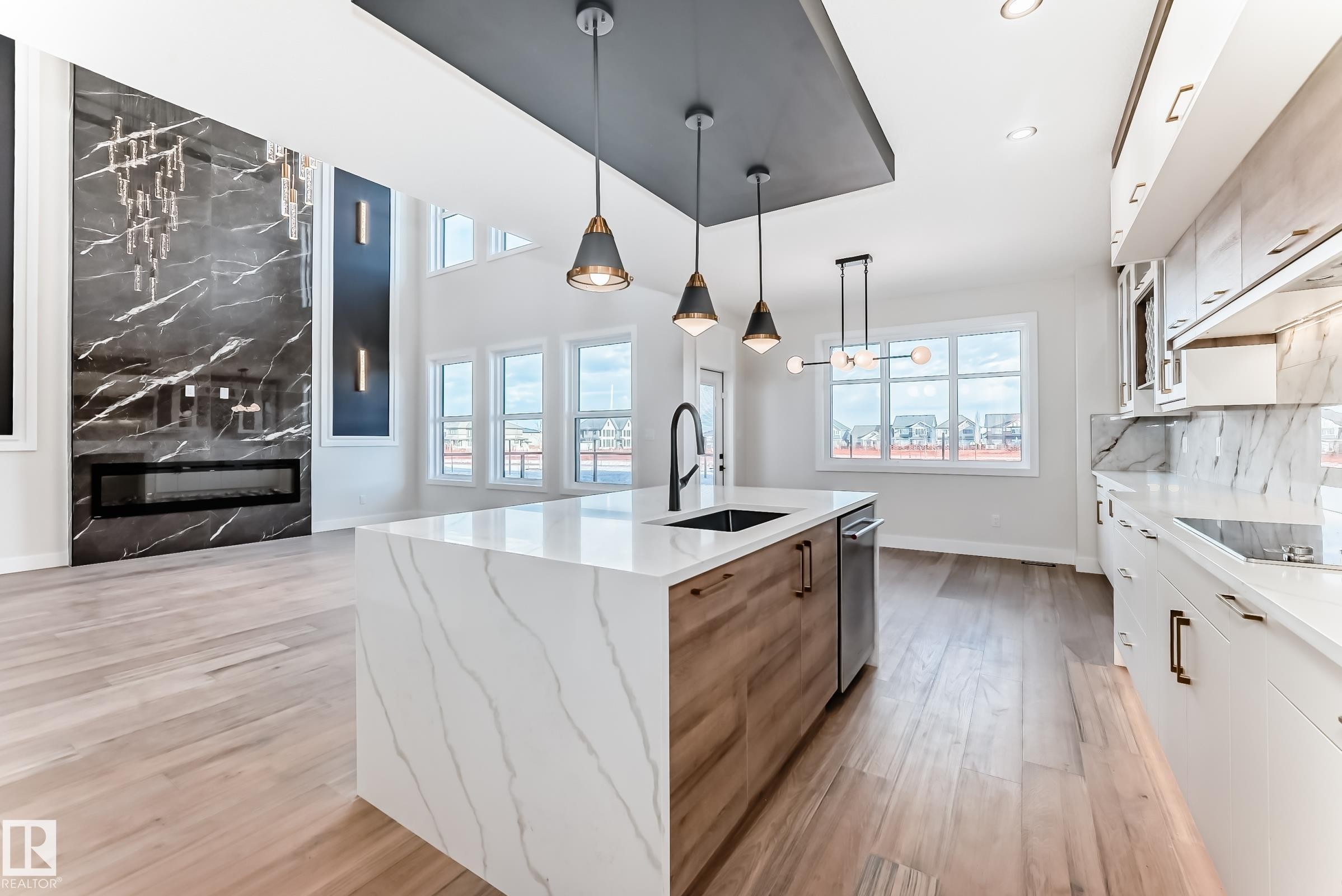 This expansive open-concept area features light hardwood flooring throughout, a kitchen island with a white marble countertop and integrated sink, and an elegant fireplace with a dark marble surround - 7665 Kimiwan Crescent, Edmonton, AB - Indoor Photo Showing Kitchen With Upgraded Kitchen