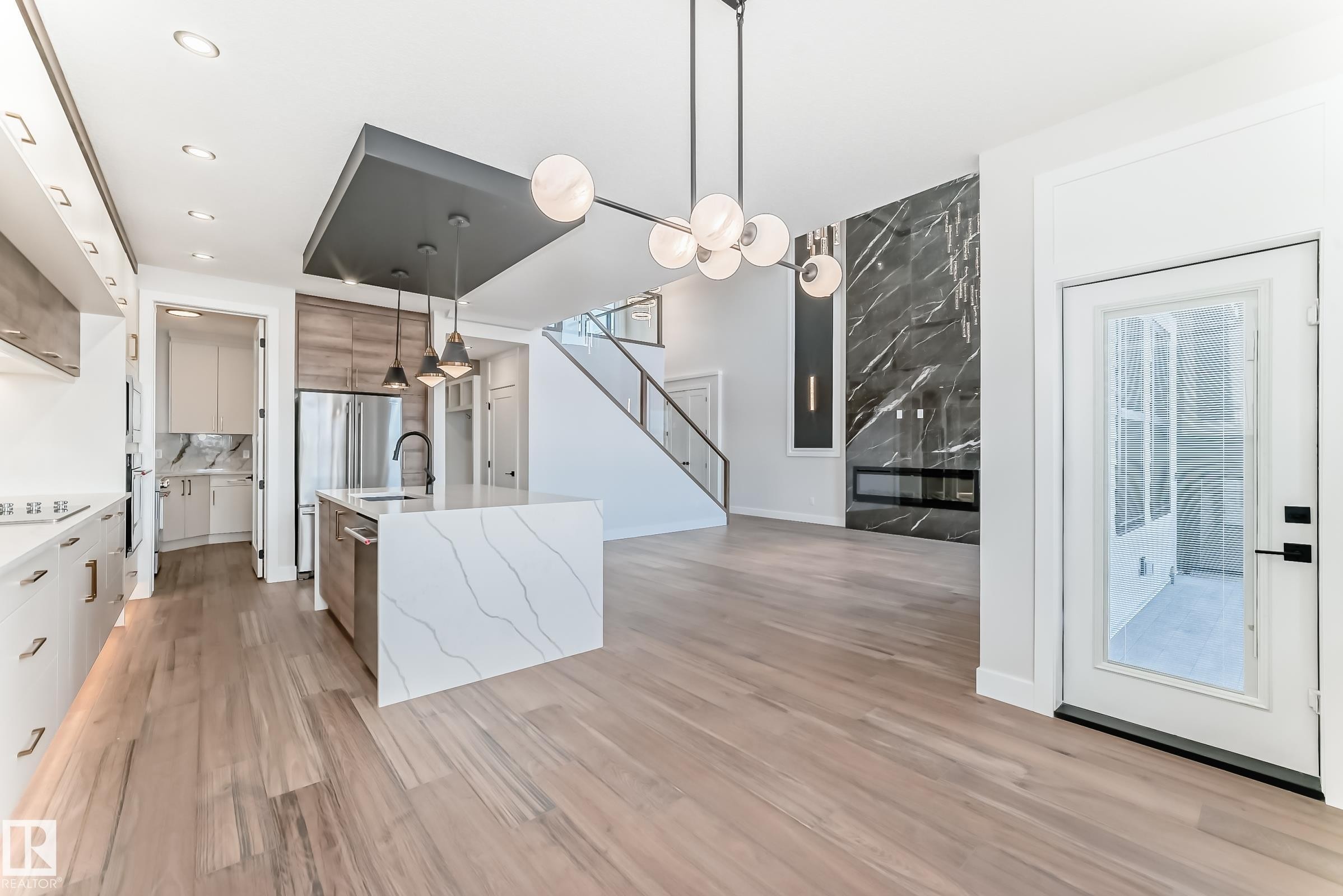 This open-concept property features hardwood flooring, a modern kitchen island with an integrated sink and pendant lighting, and a striking black marble fireplace - 7665 Kimiwan Crescent, Edmonton, AB - Indoor Photo Showing Kitchen With Upgraded Kitchen