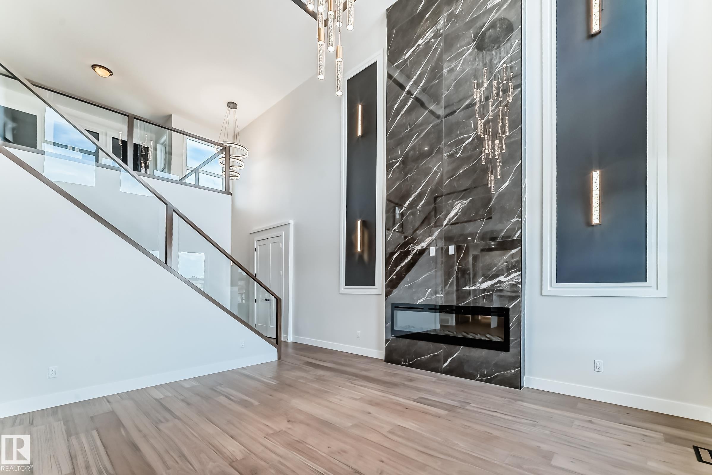 Living space featuring light hardwood flooring, a modern fireplace with a dark marble-patterned surround, a glass-paneled staircase, and tall ceilings with a chandelier - 7665 Kimiwan Crescent, Edmonton, AB - Indoor With Fireplace