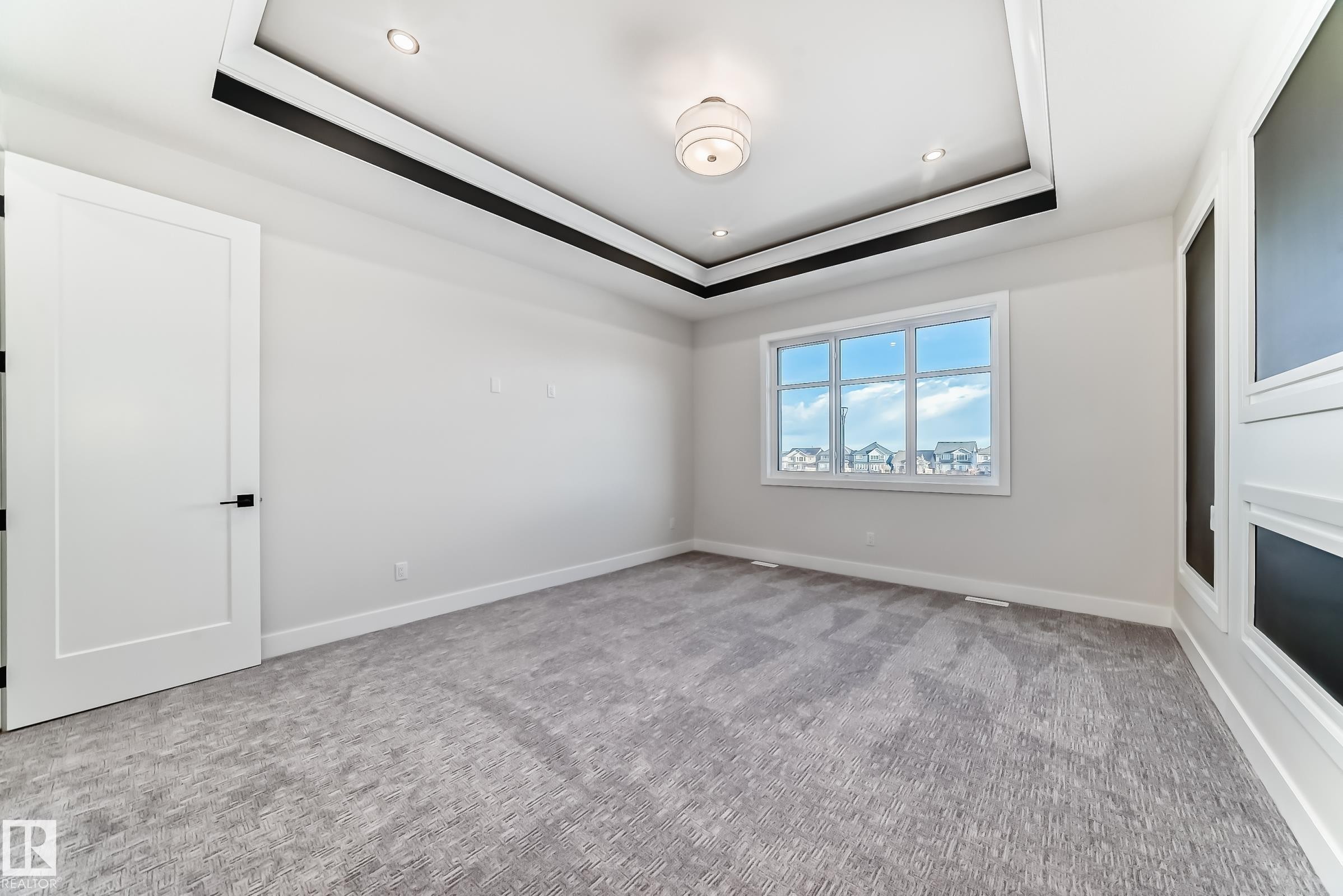 This room features a tray ceiling with recessed lighting and a central light fixture, a large window, a white door with black hardware, and patterned gray carpeting - 7665 Kimiwan Crescent, Edmonton, AB - Indoor Photo Showing Other Room