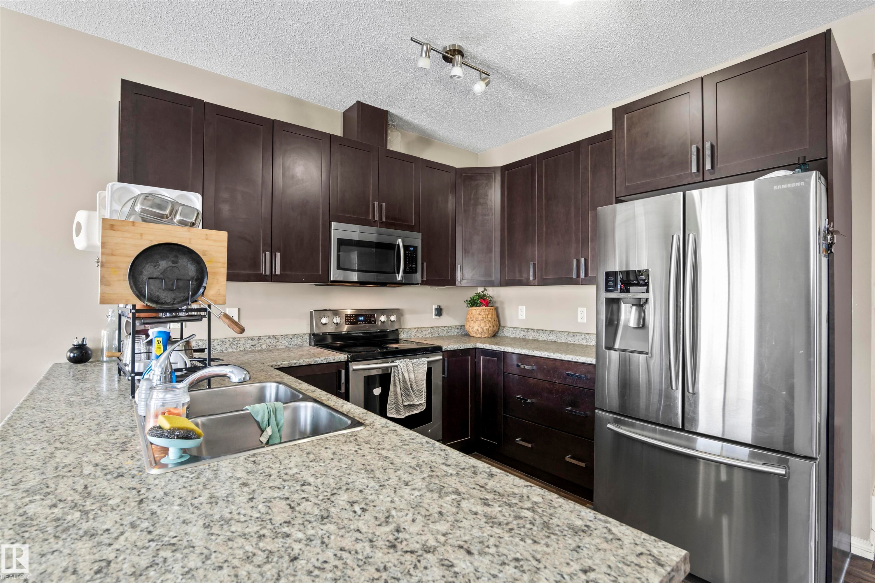 Kitchen with appliances with stainless steel finishes, dark brown cabinets, light stone countertops, a textured ceiling, and rail lighting - 2526 19A Avenue Nw, Edmonton, AB - Indoor Photo Showing Kitchen With Stainless Steel Kitchen With Double Sink With Upgraded Kitchen