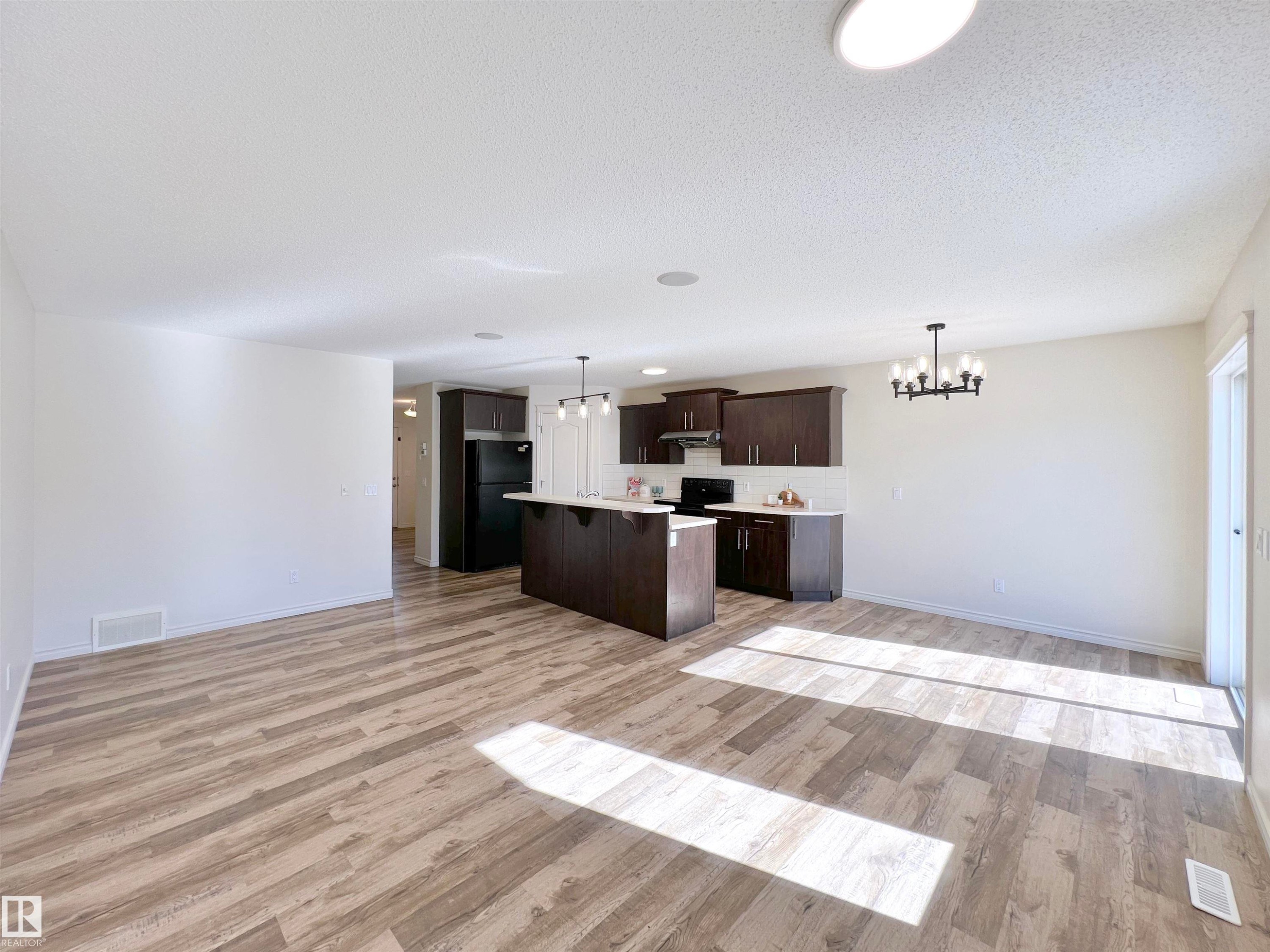 Open-concept living space featuring wood-look flooring, a kitchen with dark cabinetry, an island with seating, and a modern chandelier - 17127 7A Avenue, Edmonton, AB - Indoor Photo Showing Kitchen