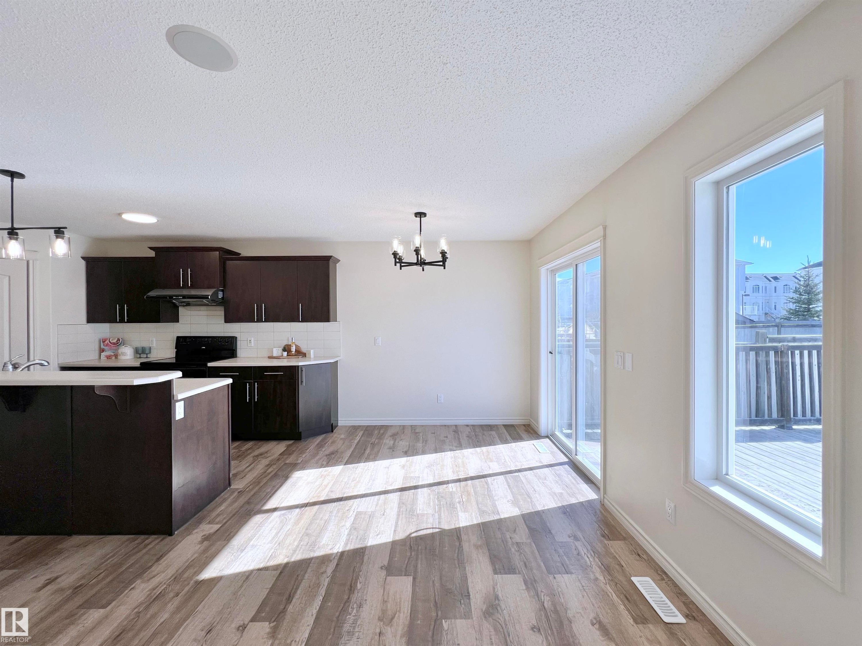 This open-concept area features wood-look flooring, a kitchen with dark cabinetry, and white subway tile backsplash - 17127 7A Avenue, Edmonton, AB - Indoor Photo Showing Kitchen