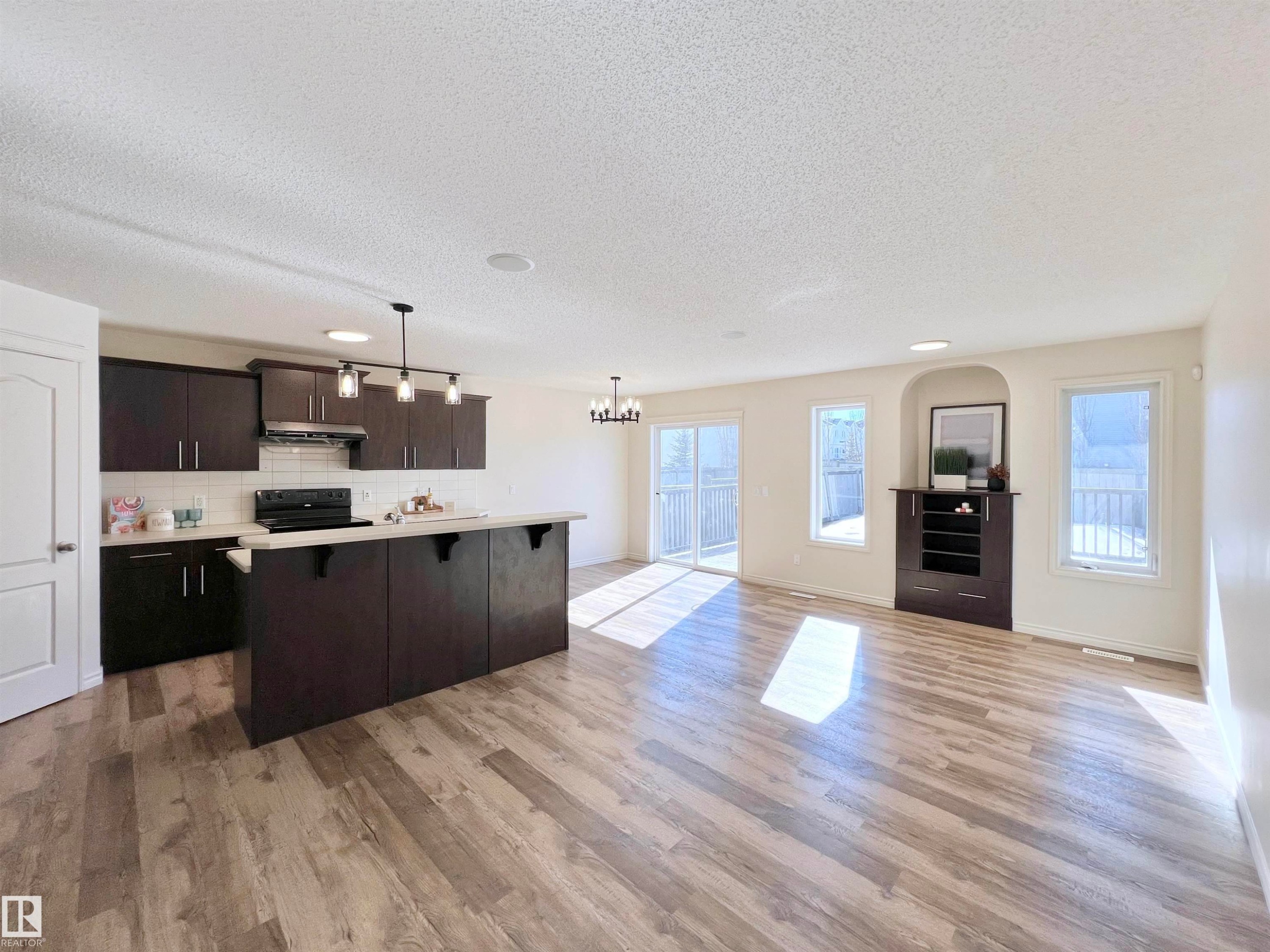 This inviting open-concept living space features light wood-look flooring, a kitchen with dark cabinetry and a central island, and sliding glass doors leading to an outdoor area - 17127 7A Avenue, Edmonton, AB - Indoor Photo Showing Kitchen