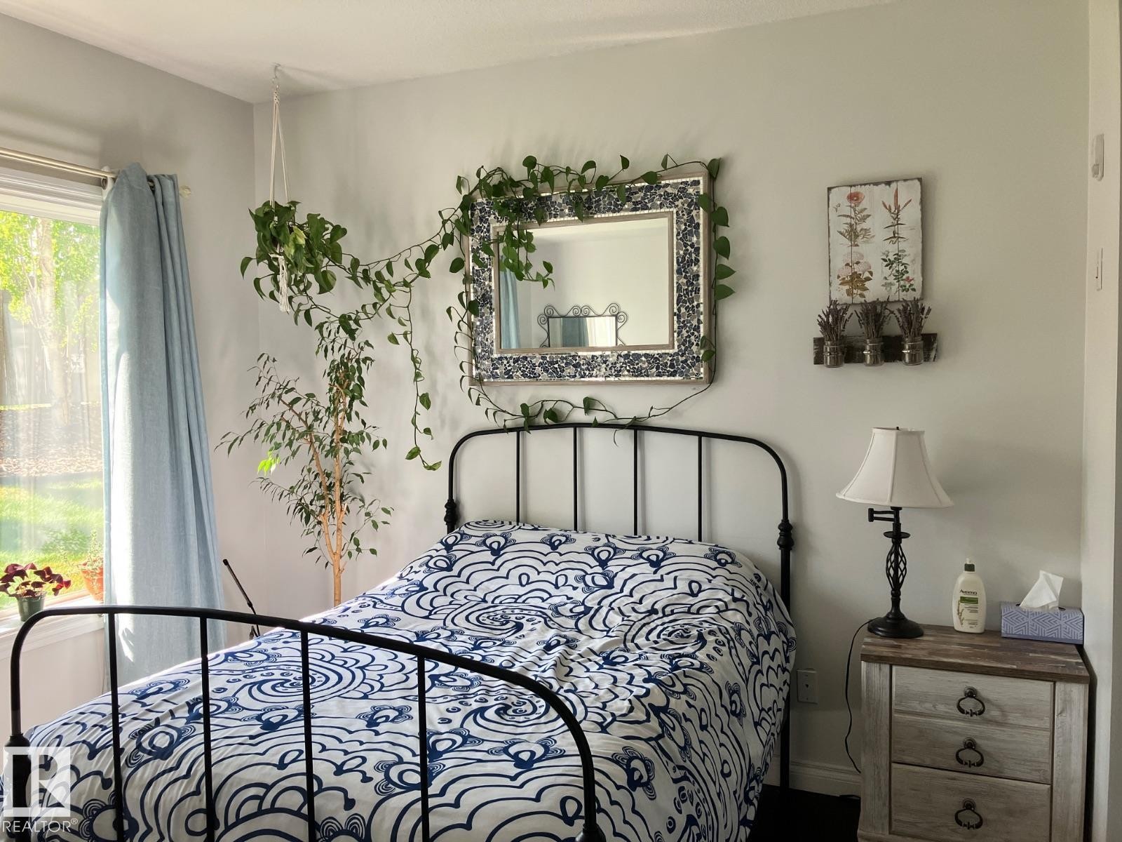 This bedroom features a window with light blue curtains, a decorative mirror with a patterned frame, and a rustic-style nightstand with three drawers - 145 10121 80 Avenue, Edmonton, AB - Indoor Photo Showing Bedroom