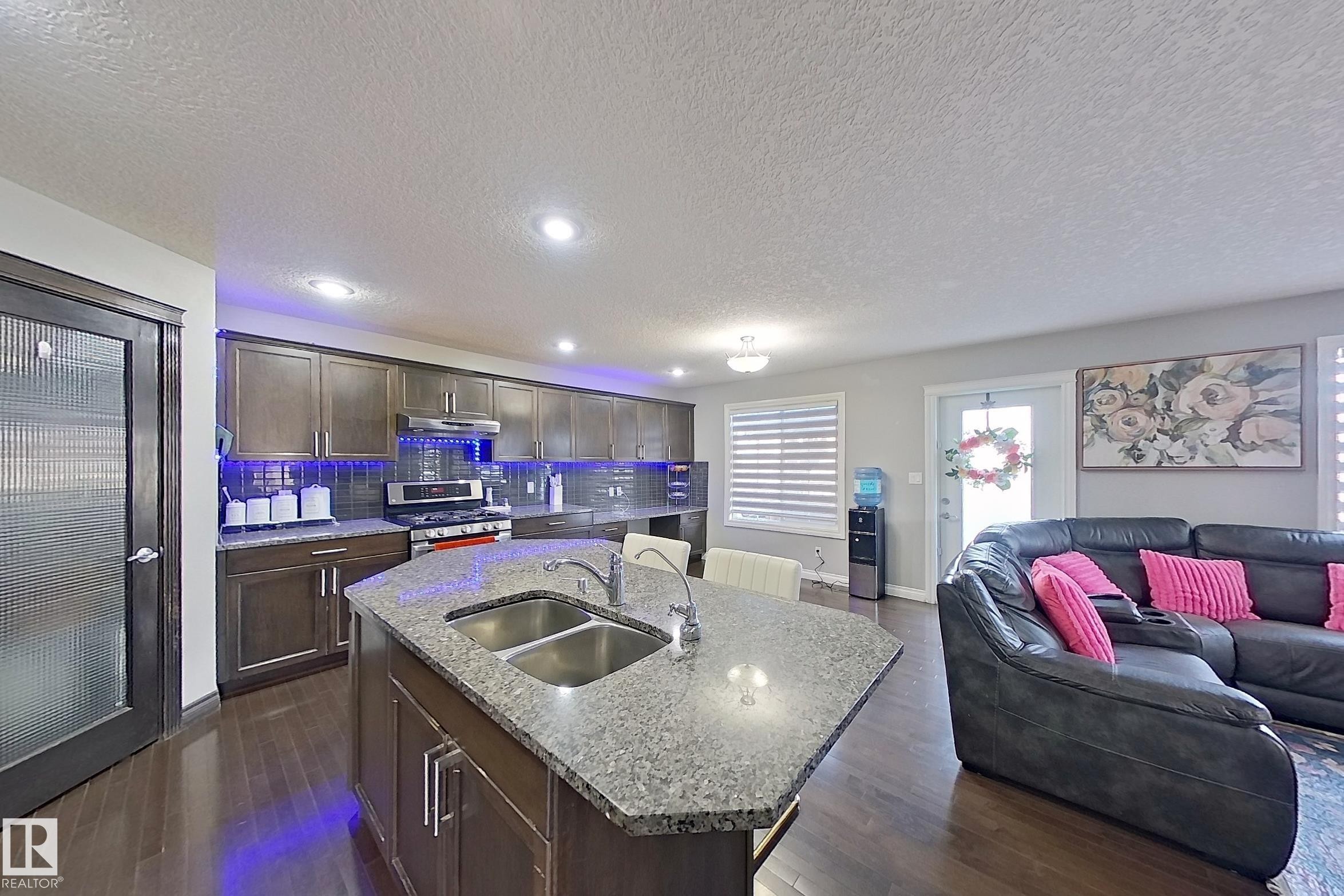 Open-concept living space with dark wood flooring, a kitchen featuring dark cabinetry, stainless steel appliances, and an island with a double sink, and a comfortable lounge area - 1743 60 Street Sw, Edmonton, AB - Indoor Photo Showing Other Room