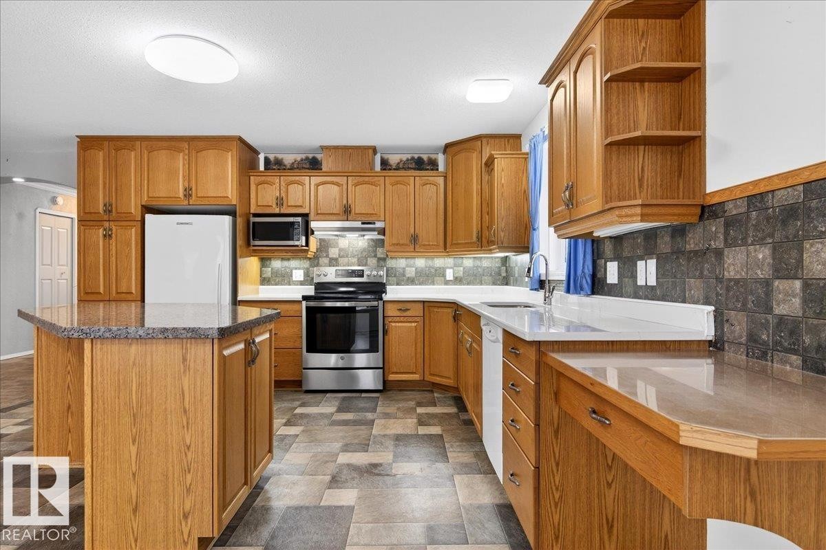 The kitchen features wood cabinetry, a central island with a countertop, stainless steel appliances including a refrigerator, oven, and microwave, and a tile backsplash - 4412 Yeoman Close, Onoway, AB - Indoor Photo Showing Kitchen With Double Sink