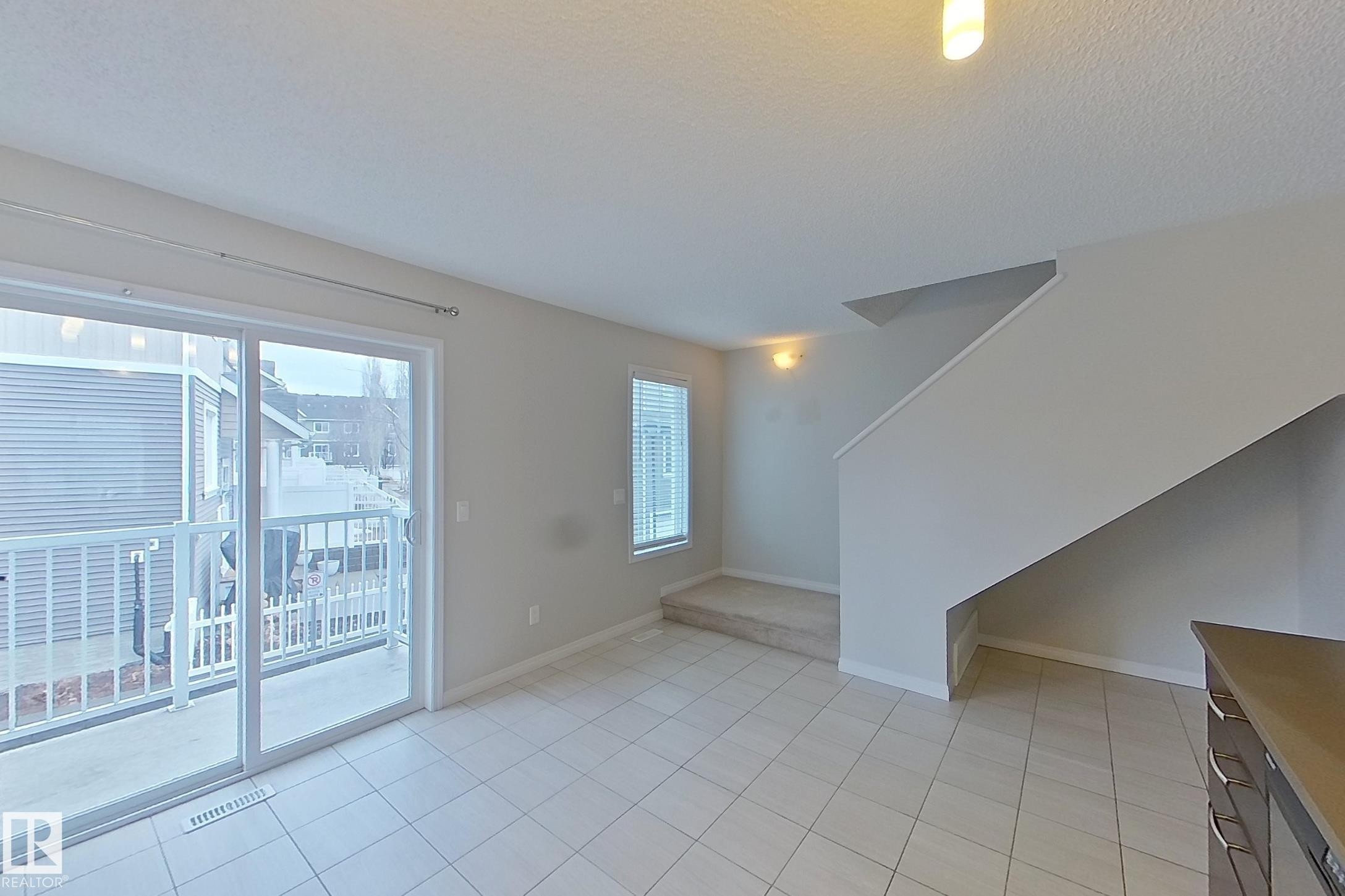 The property features a bright living area with tiled flooring, a window with blinds, and sliding glass doors leading to an outdoor balcony - 87 4050 Savaryn Drive, Edmonton, AB - Indoor Photo Showing Other Room