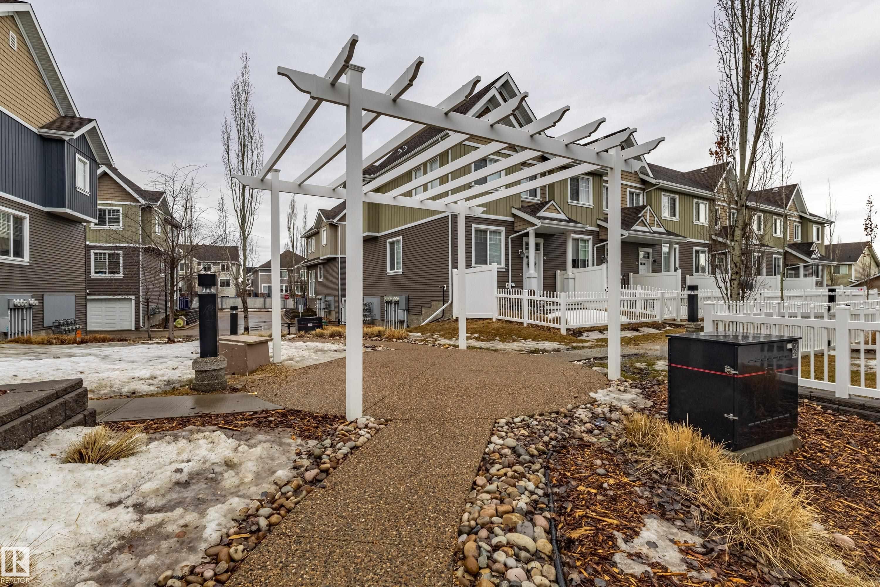 The property features an exterior with a white pergola, a paved pathway, and landscaping with decorative rocks and dry grasses - 87 4050 Savaryn Drive, Edmonton, AB - Outdoor With Facade