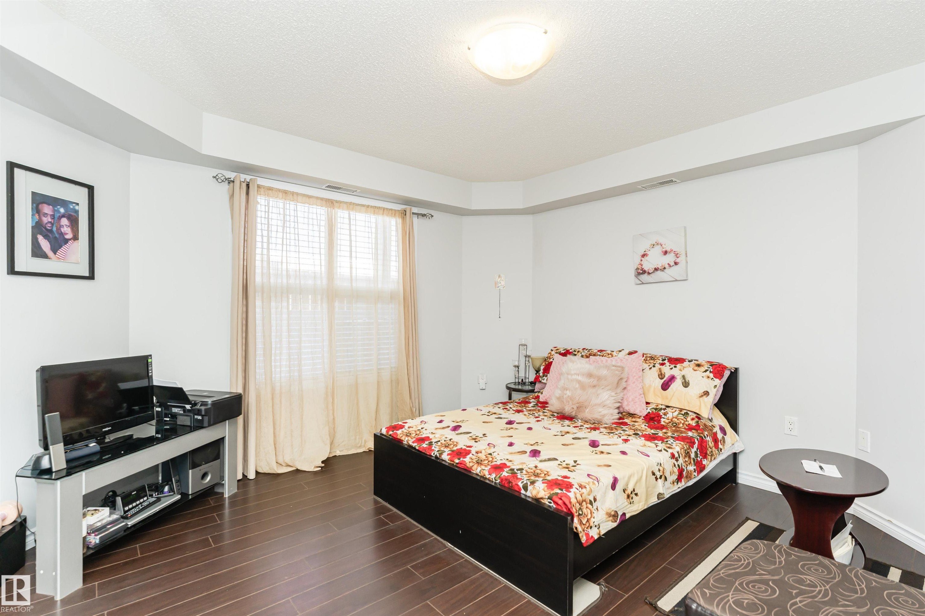 This room features dark hardwood flooring, a bright window with blinds and curtains, and a ceiling light fixture - 107 10118 106 Avenue, Edmonton, AB - Indoor Photo Showing Bedroom