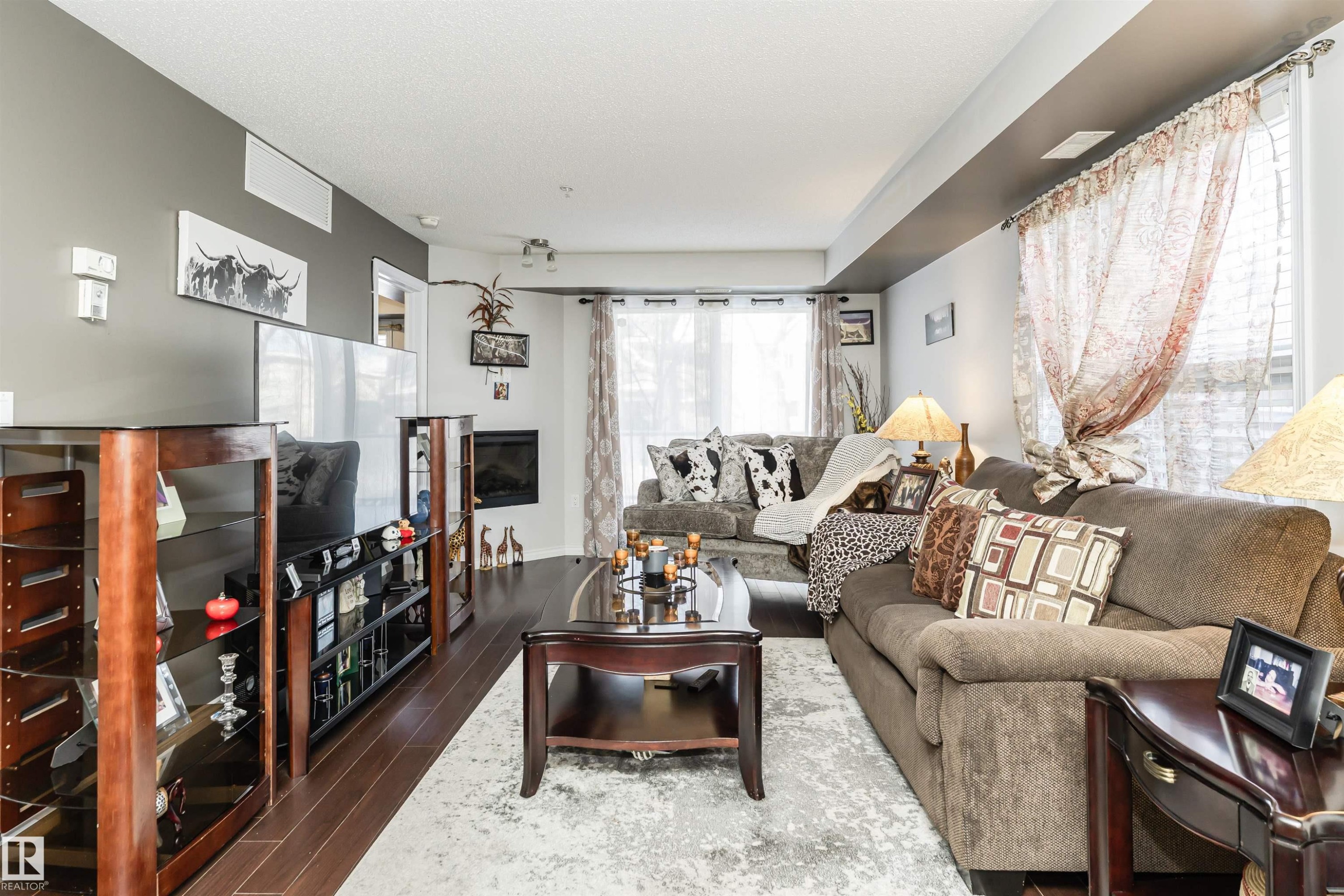 Living room featuring hardwood flooring, a fireplace, and large windows providing natural light - 107 10118 106 Avenue, Edmonton, AB - Indoor Photo Showing Living Room With Fireplace