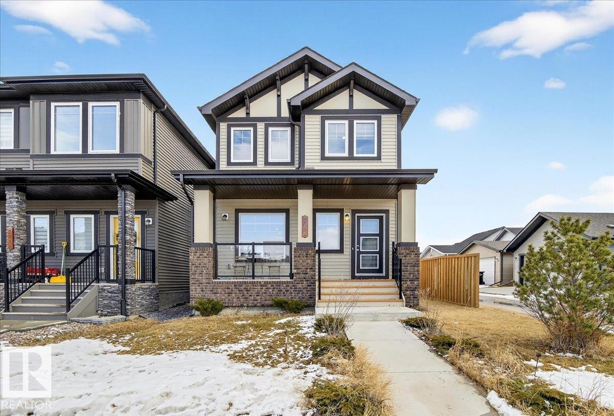 The property features a two-story exterior with light-colored siding, dark trim, and a prominent front porch - 203 Cavanagh Common, Edmonton, AB - Outdoor With Deck Patio Veranda With Facade