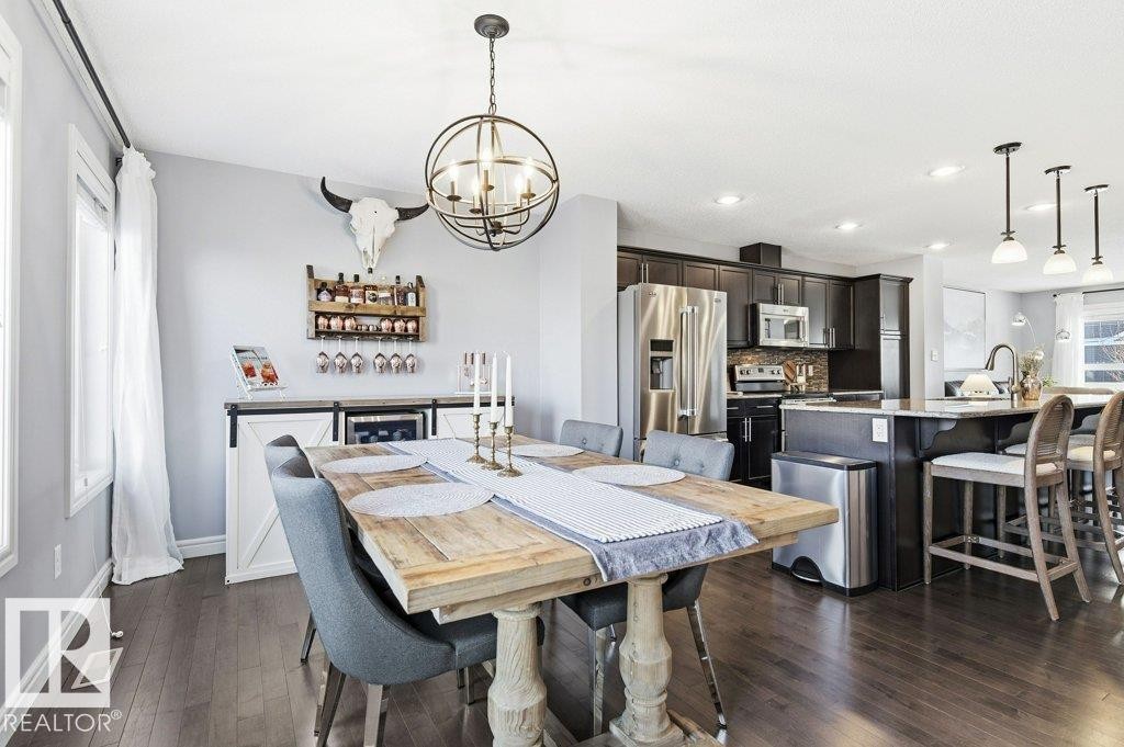Open concept living space featuring dark hardwood flooring throughout, a dining area with a rustic-style table, and a kitchen with dark cabinetry, stainless steel appliances, and an island with seating - 16720 15 Avenue, Edmonton, AB - Indoor Photo Showing Dining Room