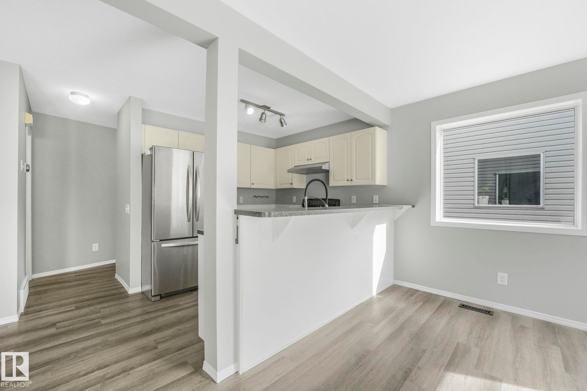 The property offers a kitchen with light-colored cabinetry, a stainless steel refrigerator, and a gray countertop, complemented by light gray walls and wood-patterned flooring - 123 230 Edwards Drive, Edmonton, AB - Indoor Photo Showing Kitchen