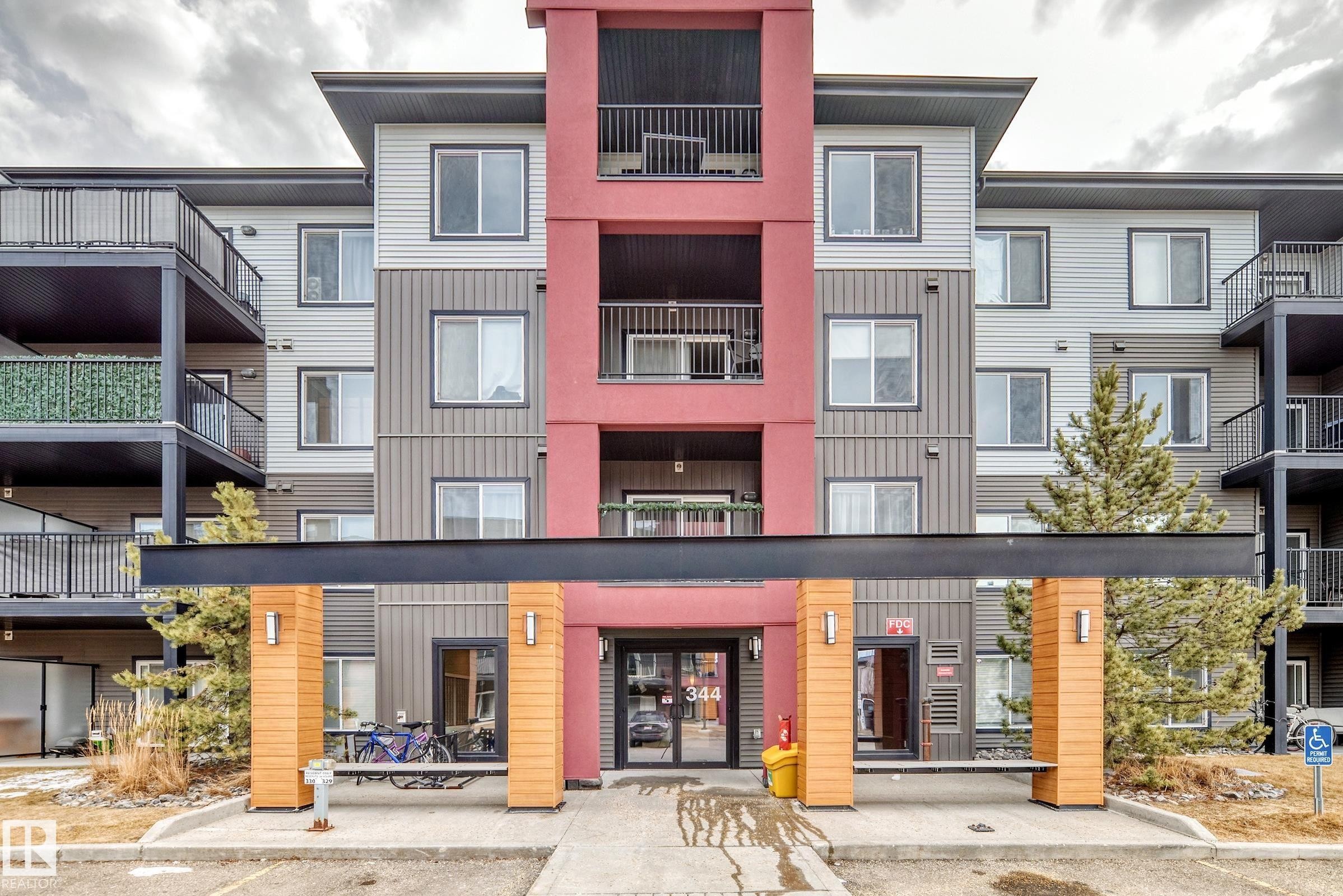 The property features a multi-story exterior with a modern design, incorporating grey and red panels, and numerous balconies with black railings - 332 344 Windermere Road, Edmonton, AB - Outdoor With Balcony With Facade