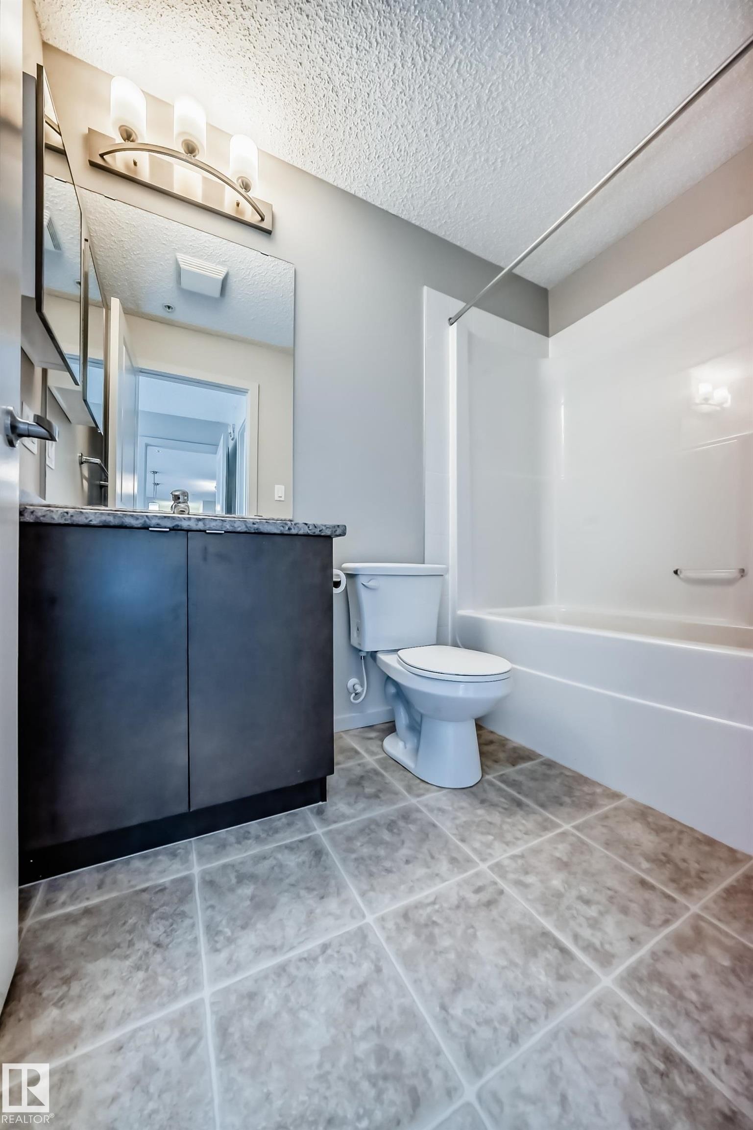 Bathroom featuring a vanity with dark cabinetry and a light-colored countertop, a toilet, and a white bathtub with a shower rod - 332 344 Windermere Road, Edmonton, AB - Indoor Photo Showing Bathroom