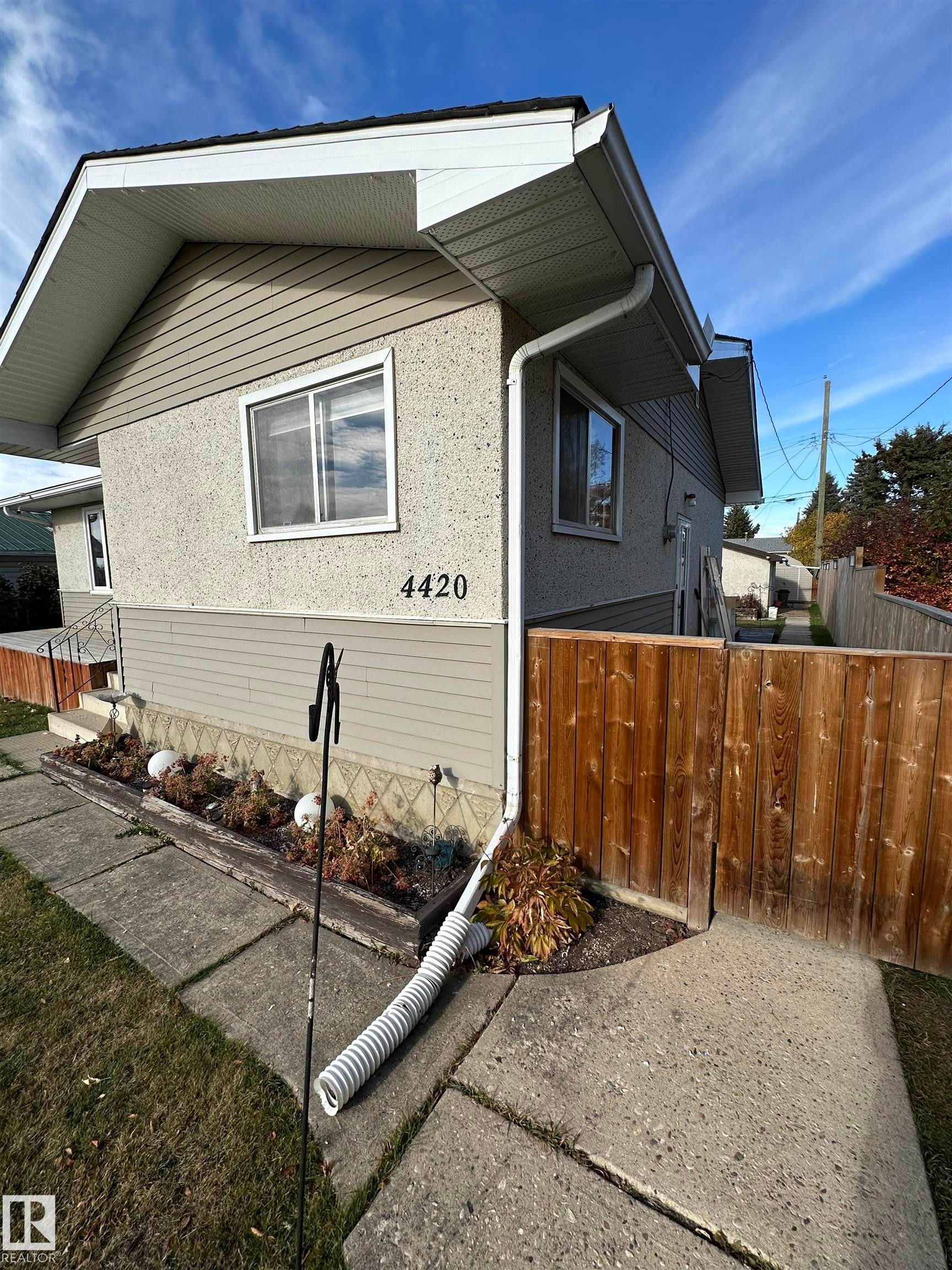 The property features a stucco and siding exterior, with white window frames, and a wooden fence - 4420 56 Avenue, Barrhead, AB - Outdoor With Exterior