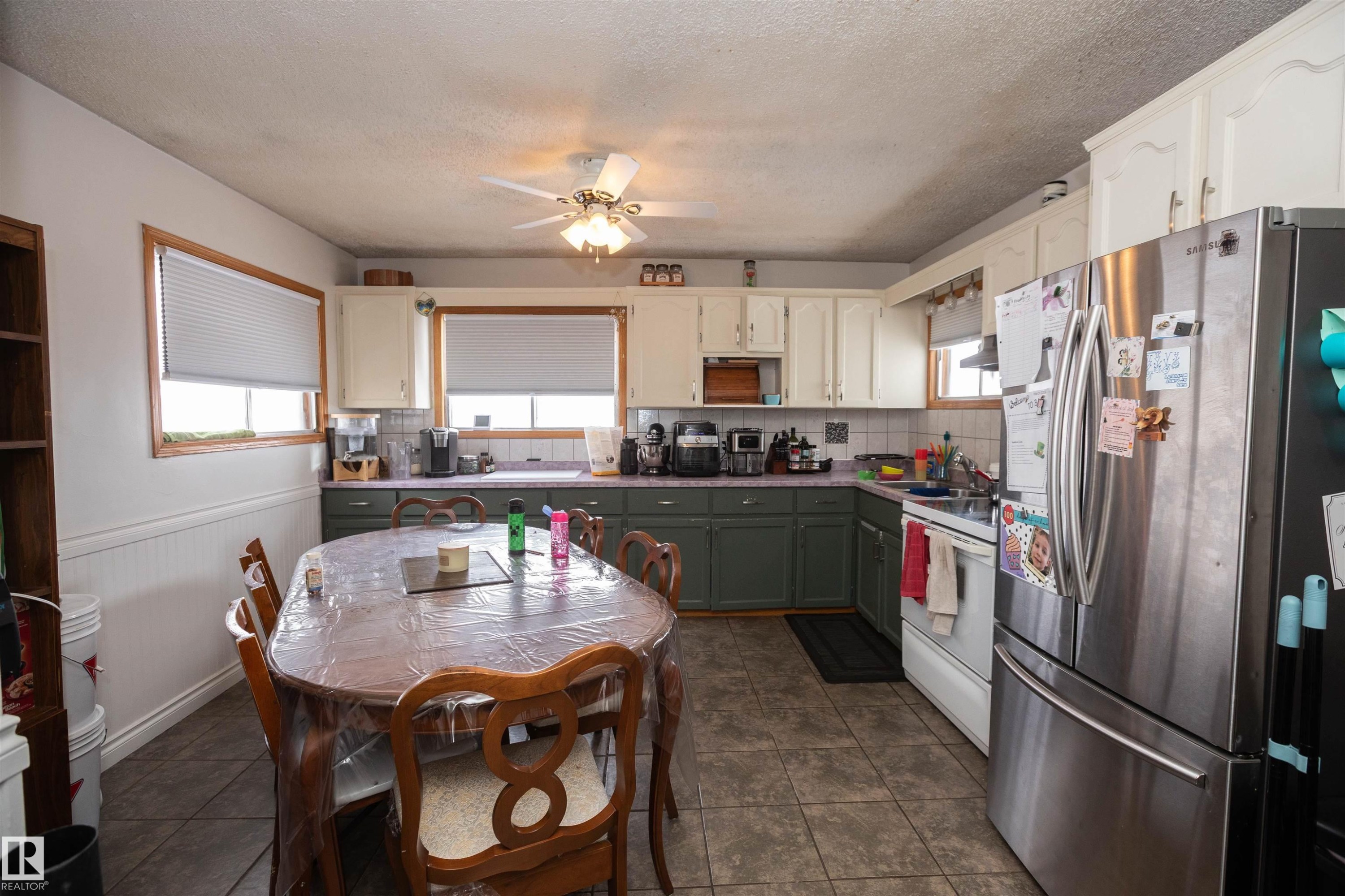 The kitchen features a dining area with a wooden table, tiled flooring, and windows with blinds - 4420 56 Avenue, Barrhead, AB - Indoor